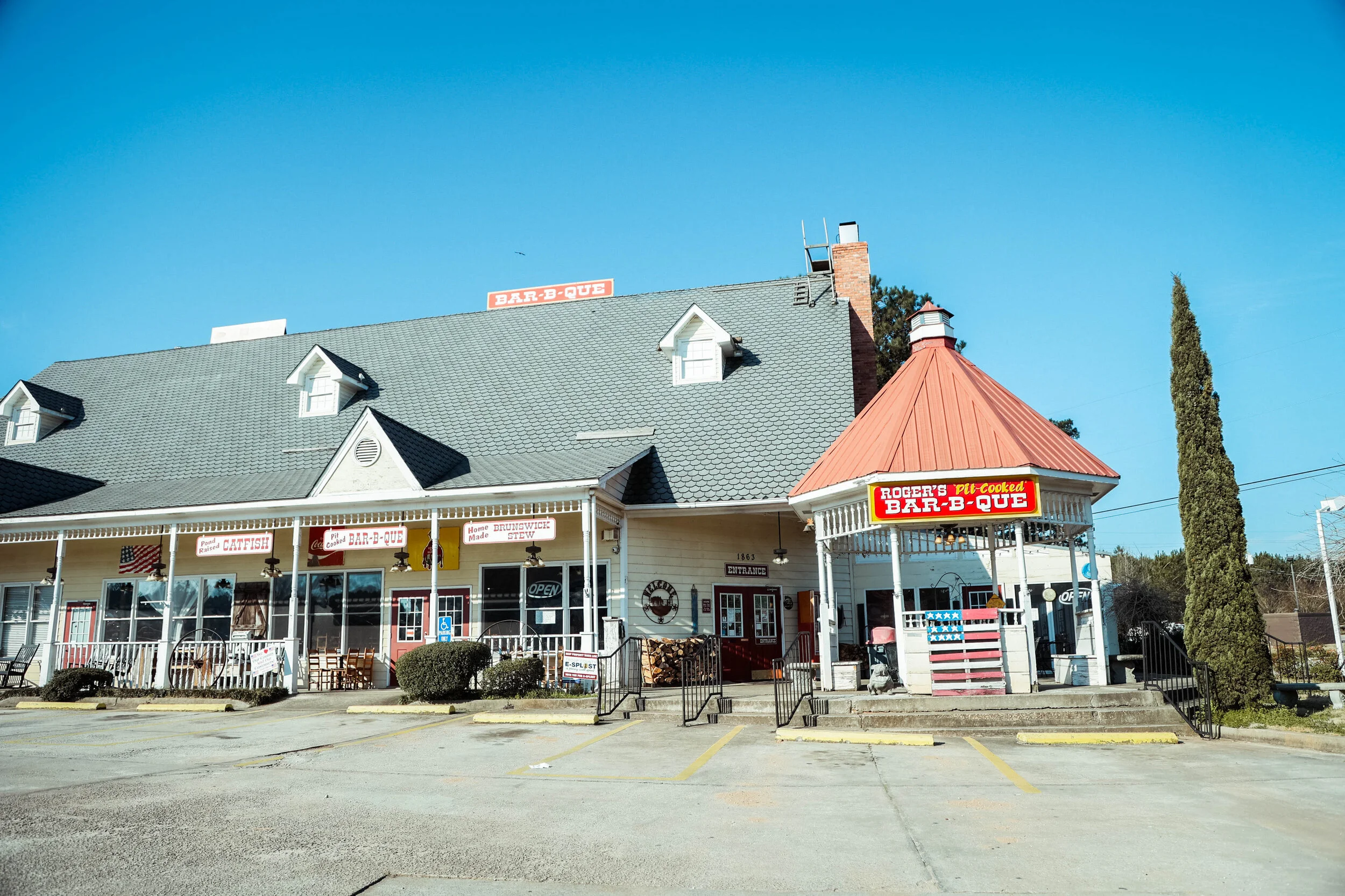 Exterior view of Roger's Bar-B-Que restaurant with signs indicating smoked catfish, home-cooked stew, and pit-cooked barbecue, under a clear blue sky.