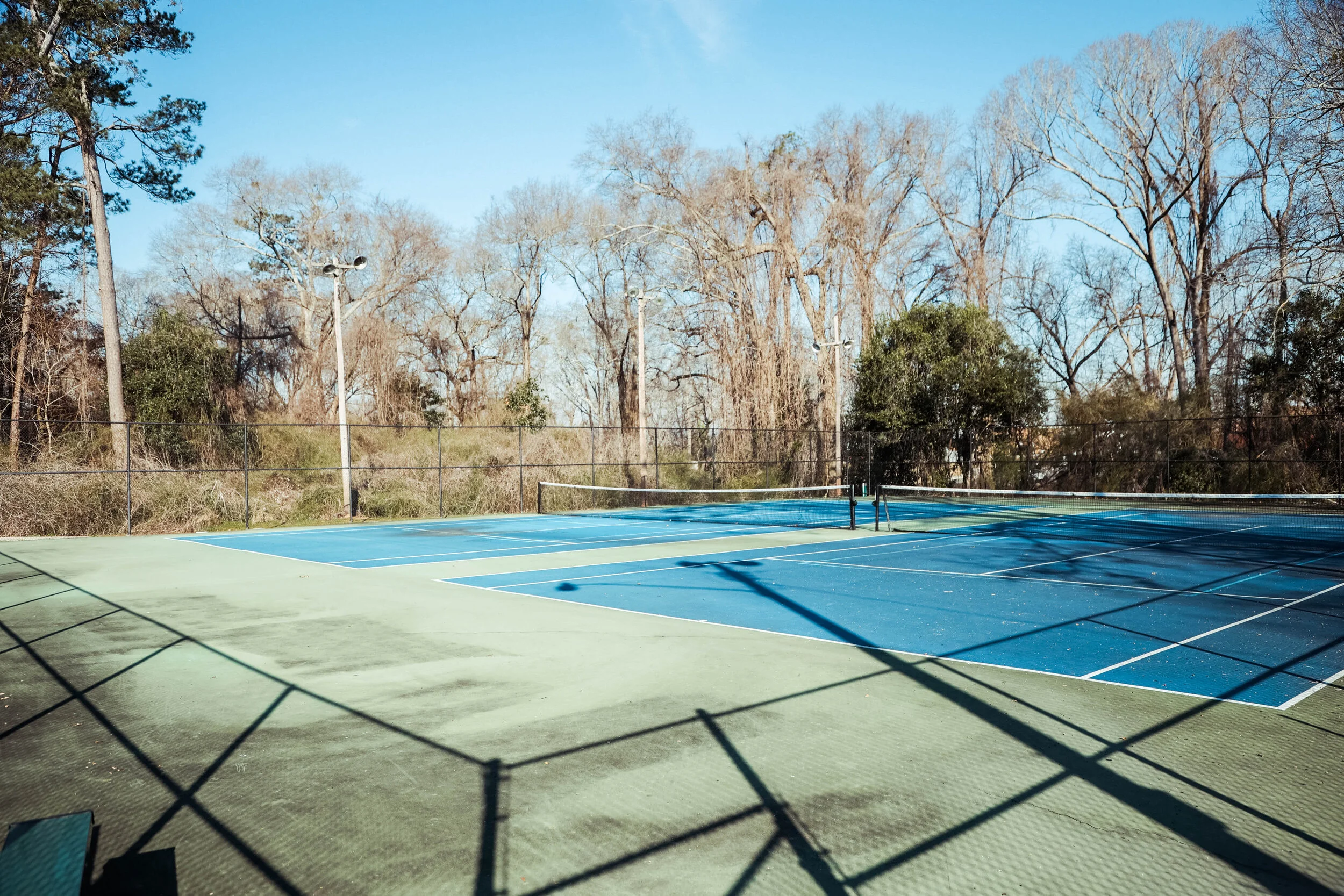 Empty outdoor tennis court with a blue surface and surrounding green area, enclosed by a chain-link fence and tall trees in the background under a clear sky.