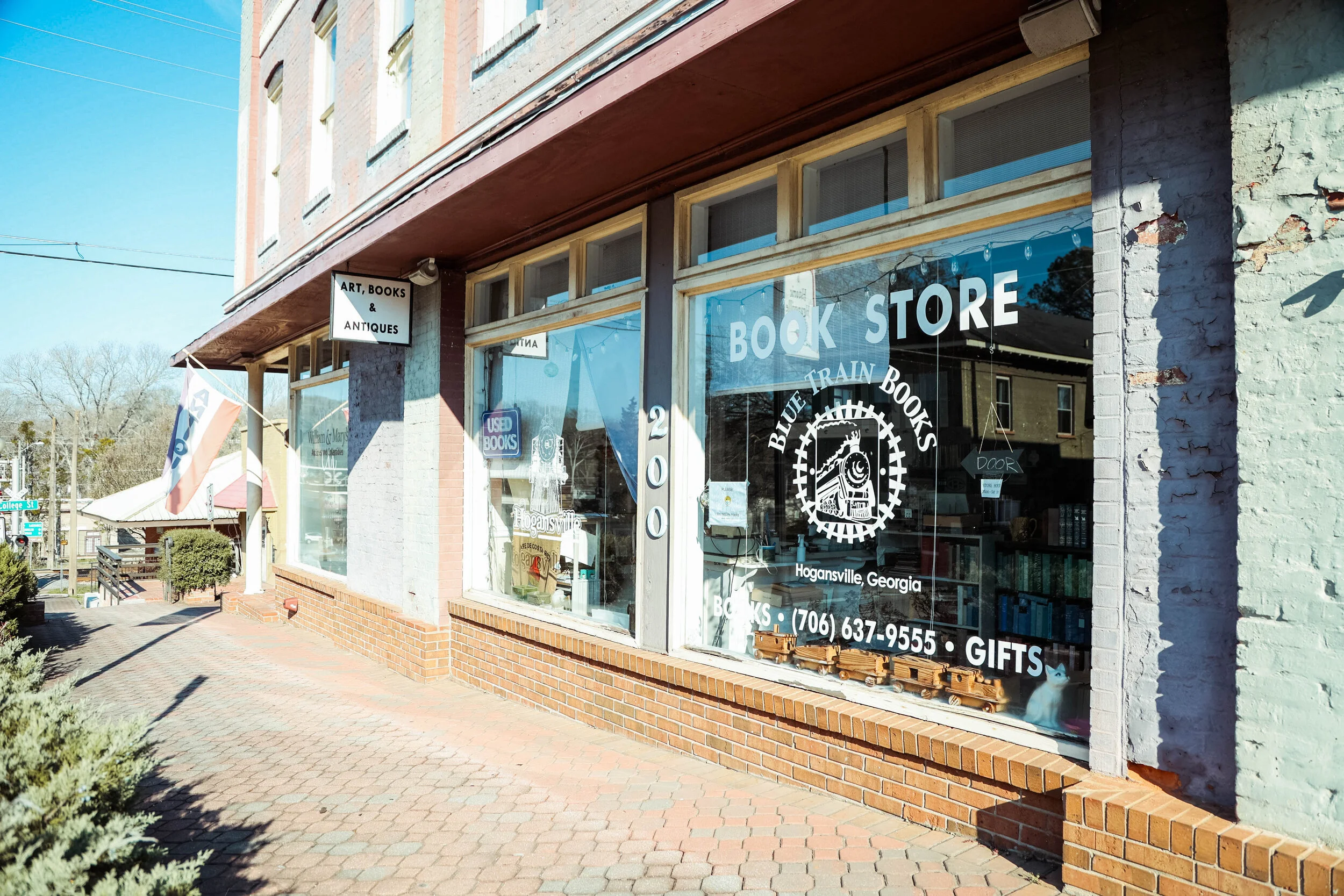 Front view of a bookstore named Blue Train Books in Hogansville, Georgia, with large windows displaying books and signs, a brick sidewalk outside, and a sign indicating it sells art, books, antiques, and gifts.