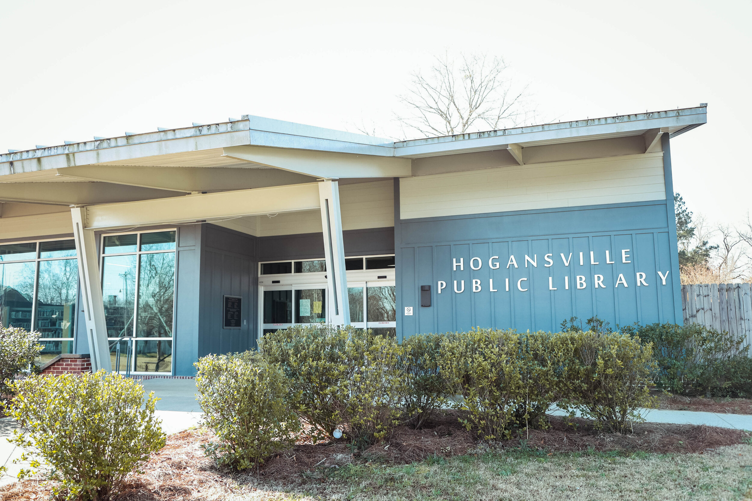 Exterior view of Hogansville Public Library building with blue siding, glass windows, and a sign displaying the library's name.