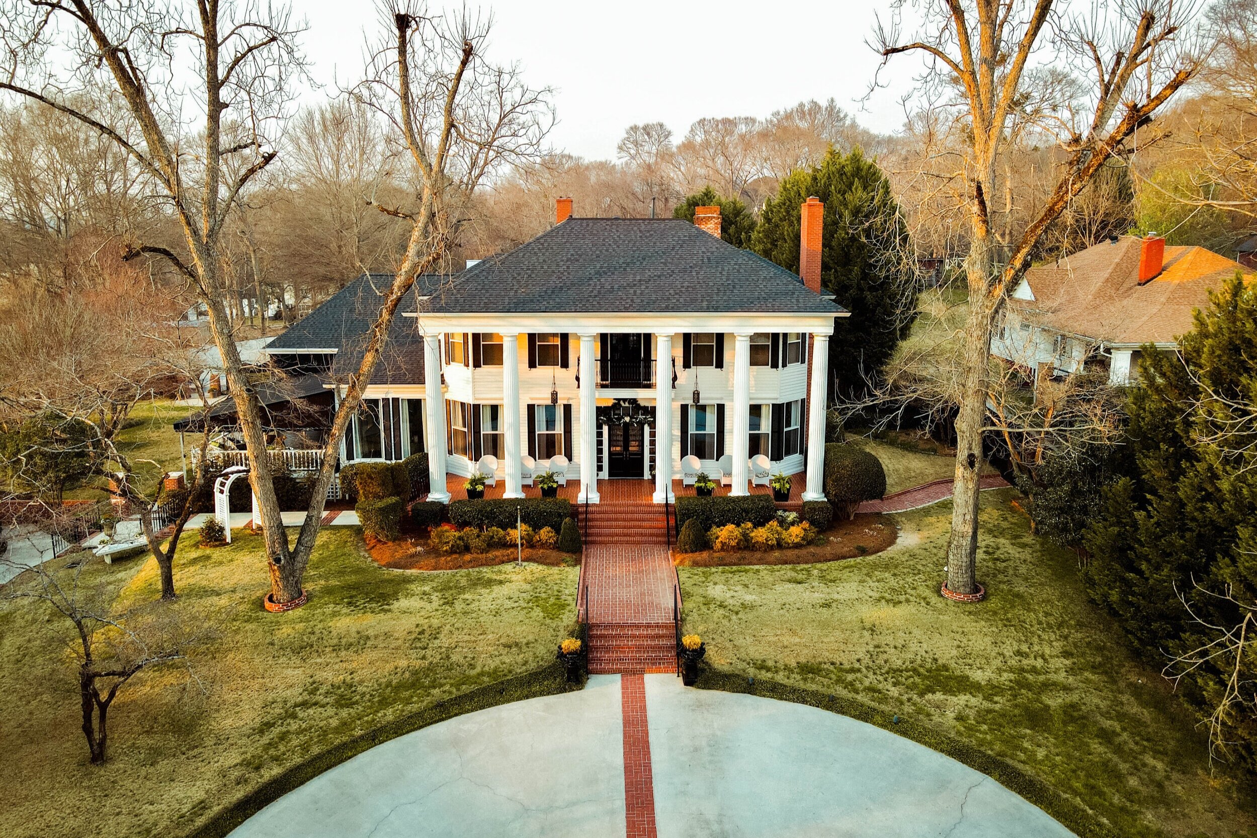 A large white house with multiple columns and black shutters, surrounded by trees and shrubs, with a brick walkway leading to the front door.
