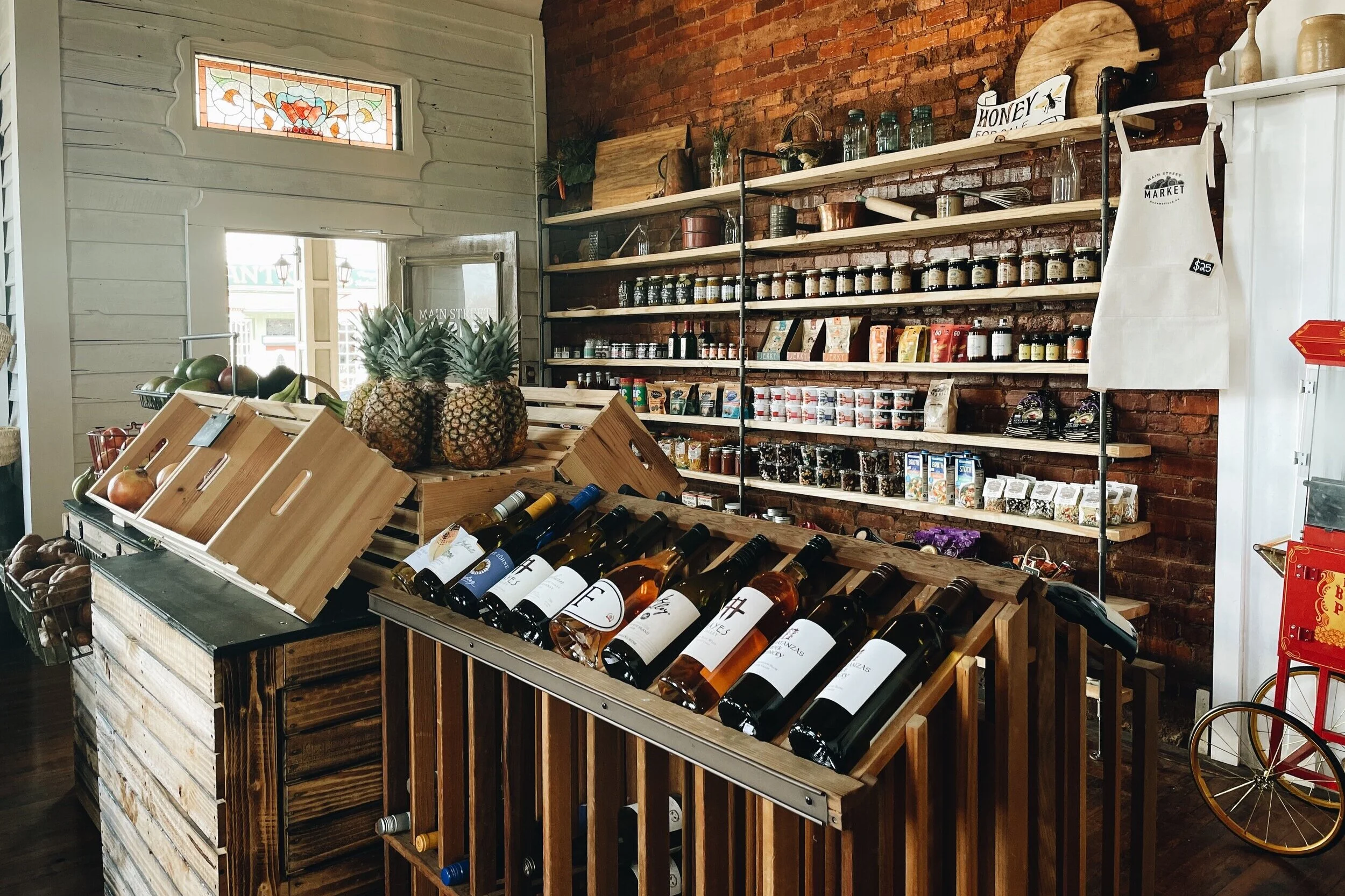 Interior of a grocery store with wooden shelves stocked with jars, bottles, and packaged goods, a display of wine bottles on a wooden rack, fresh pineapples on a crate, and other fresh produce near the window and brick wall.