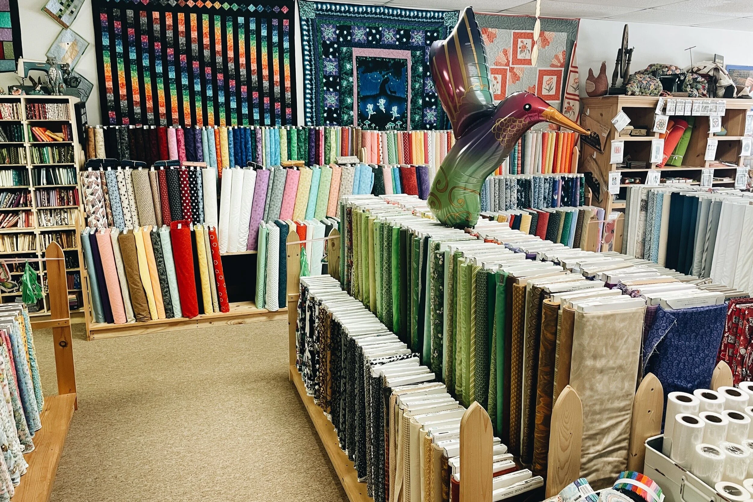 Display of multicolored fabric bolts and quilt patterns in a fabric store with a decorative inflatable bird.
