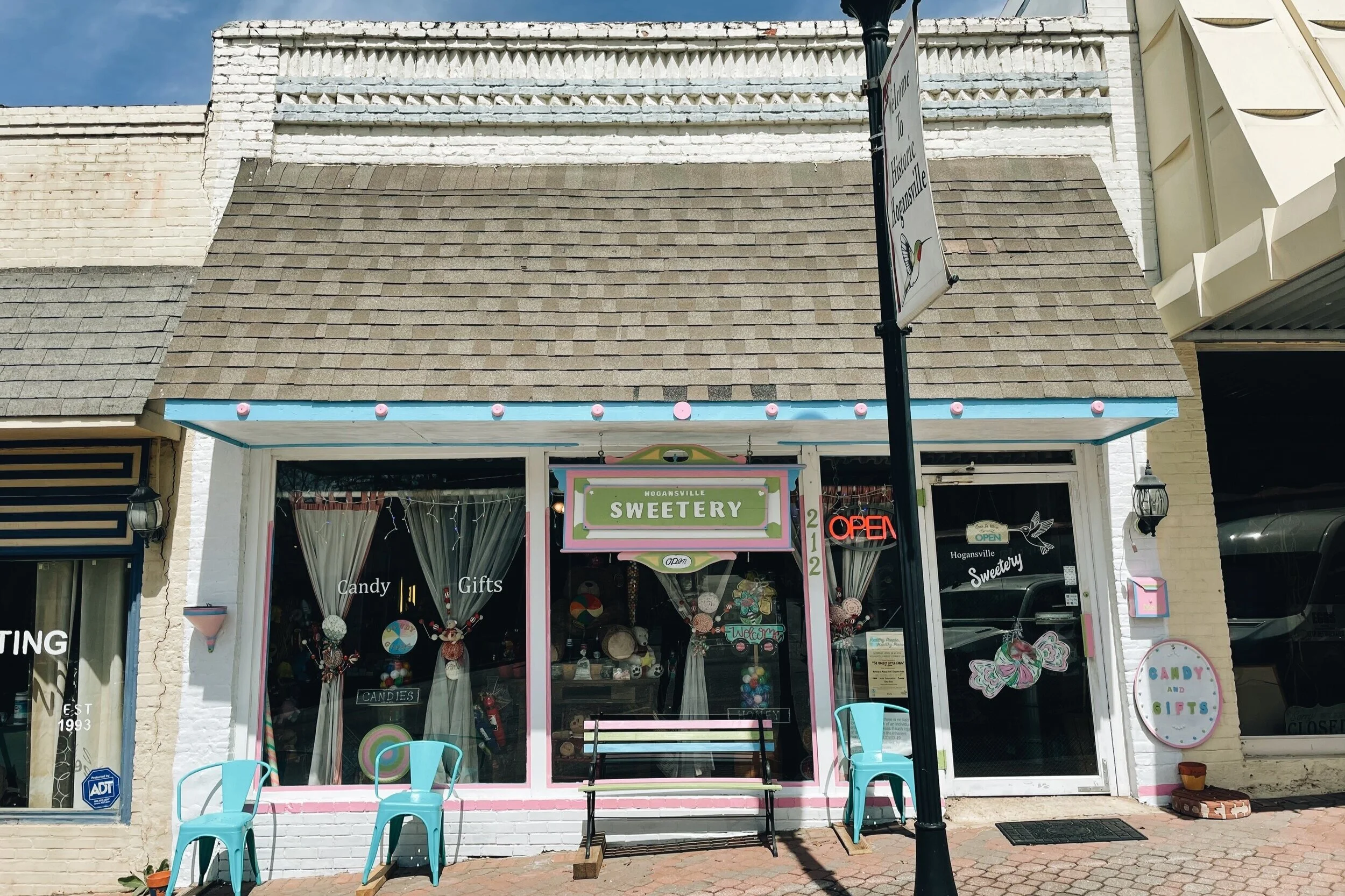 Front view of Hogansville Sweetry, a candy and gift shop, featuring a pink and white storefront with a sign above, an 'Open' neon sign, blue chairs outside, and decorative window displays with candy-themed items.