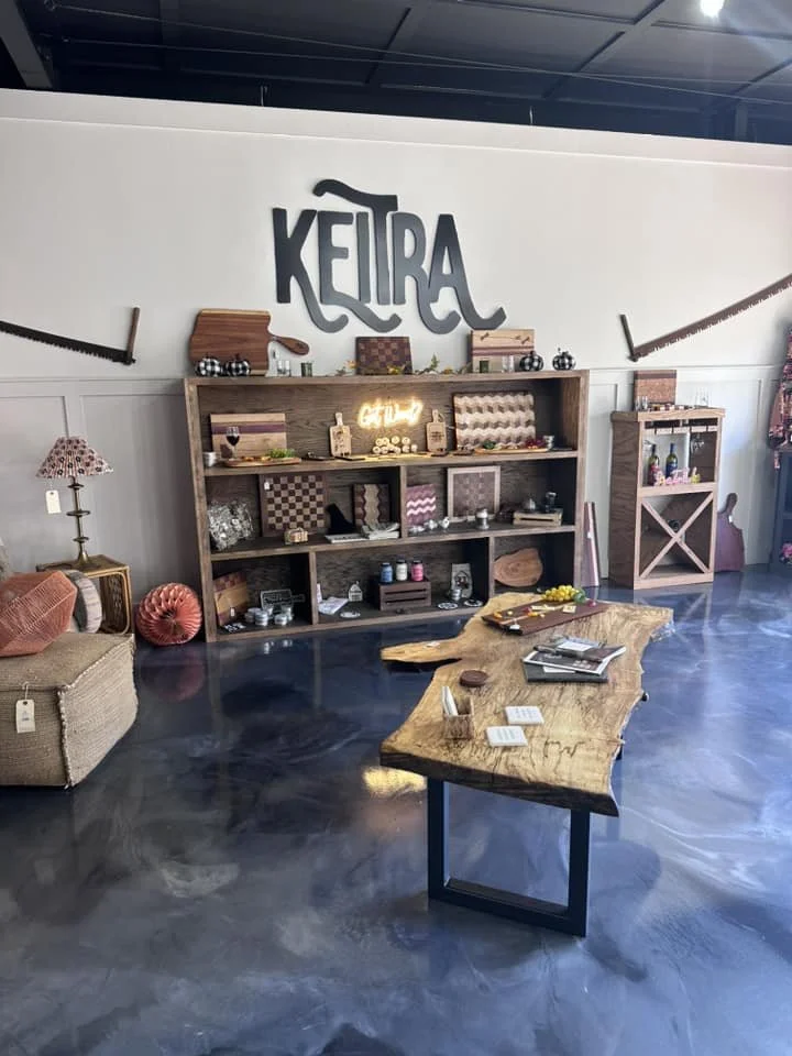 Interior of a room with a large sign that says 'KEILRA' on the wall, decorated with rustic furniture, including a wooden table, shelves with decorative items, a wicker armchair, and various decorations like lamps, paintings, and small collectibles.