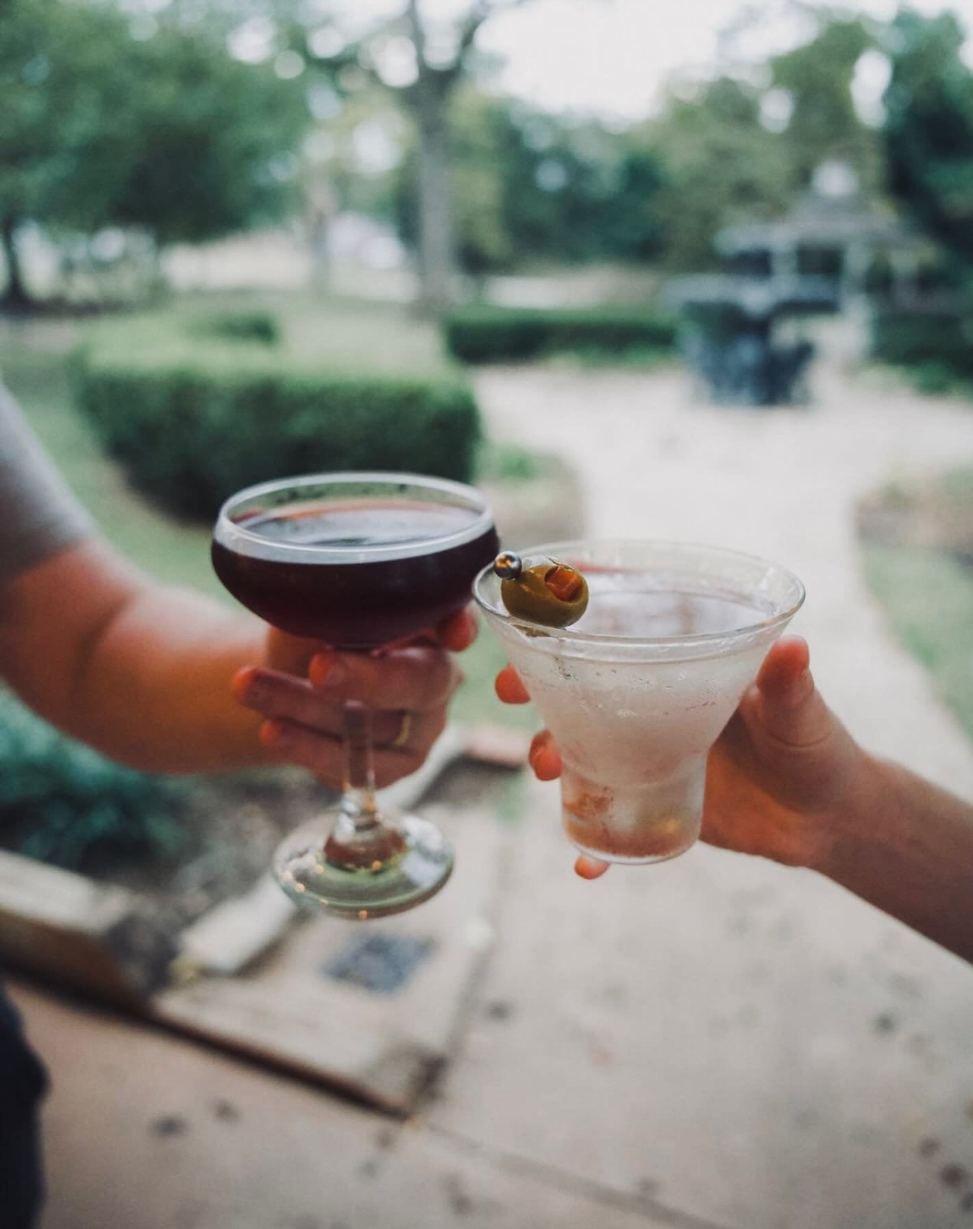 Two hands holding cocktails, one with a dark red drink in a coupe glass and the other with a clear drink garnished with an olive in a martini glass, outside in a garden setting.