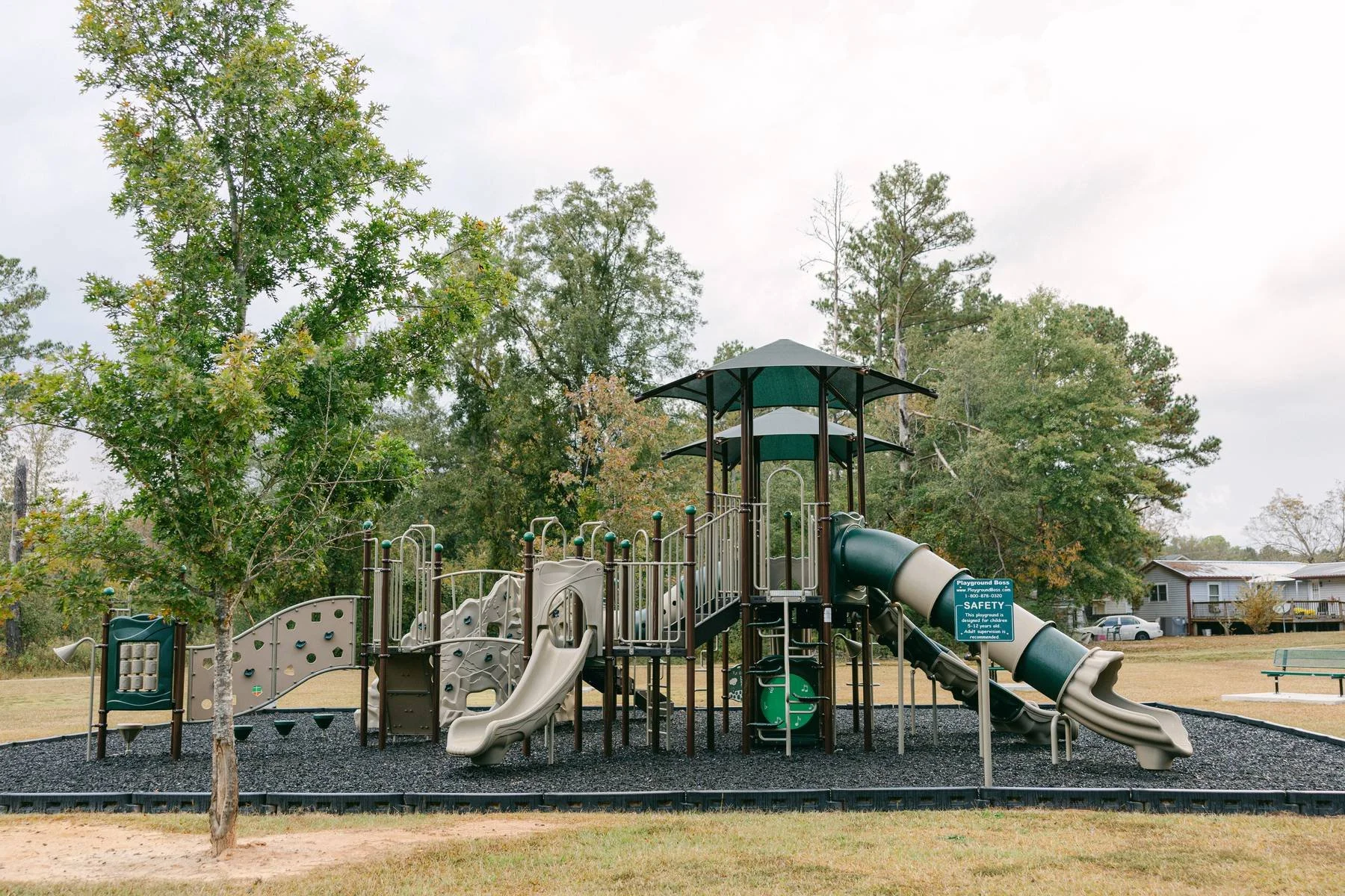 Empty playground with slides and climbing structures, surrounded by trees and residential homes, on a cloudy day.