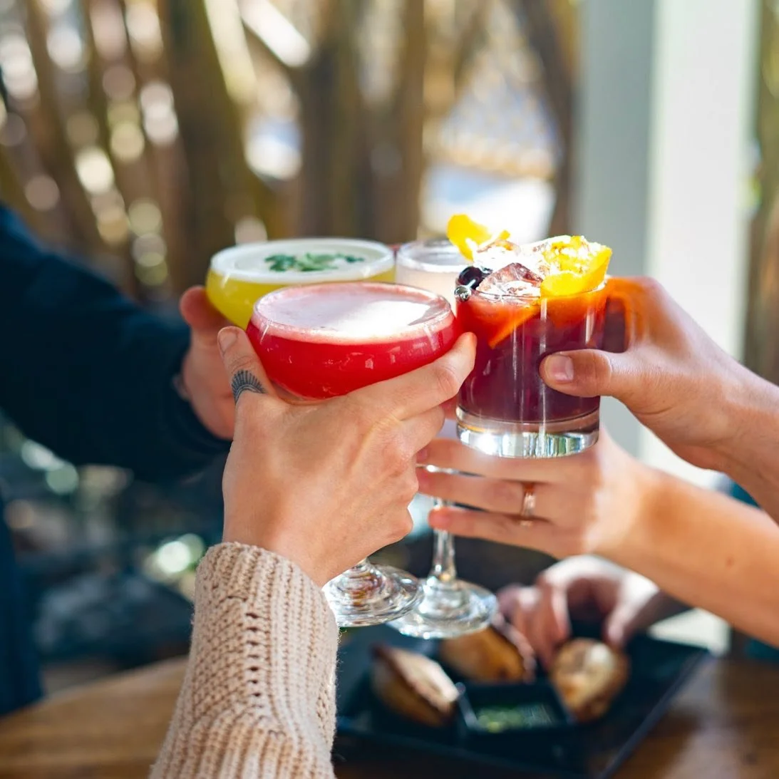 Four people holding colorful cocktails in their hands for a toast, with a plate of food in the background.
