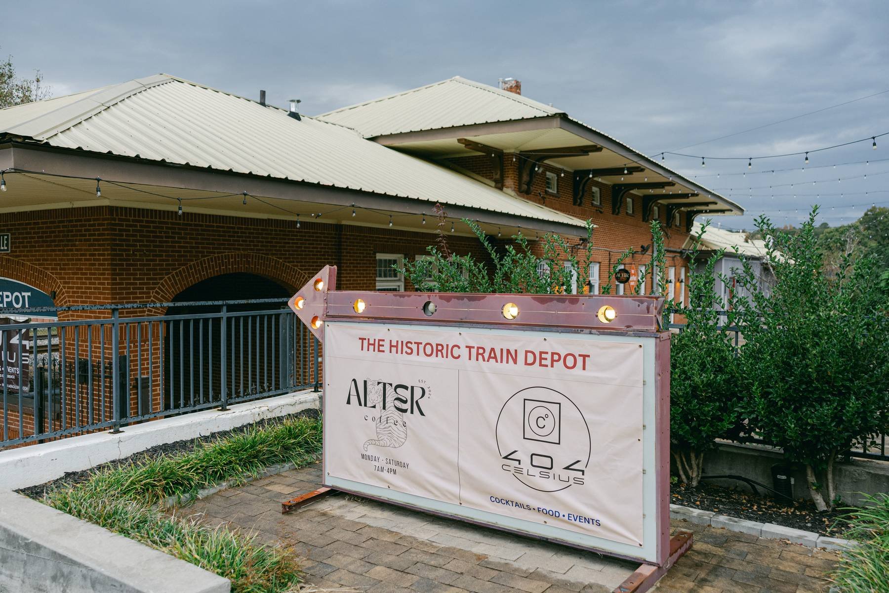 A sign outside a building reads 'The Historic Train Depot' with two logos and text for Alter Coffee and C.O. Celsius, advertising cocktails, food, and events. The building has a brown brick exterior with a light-colored metal roof. There are string l