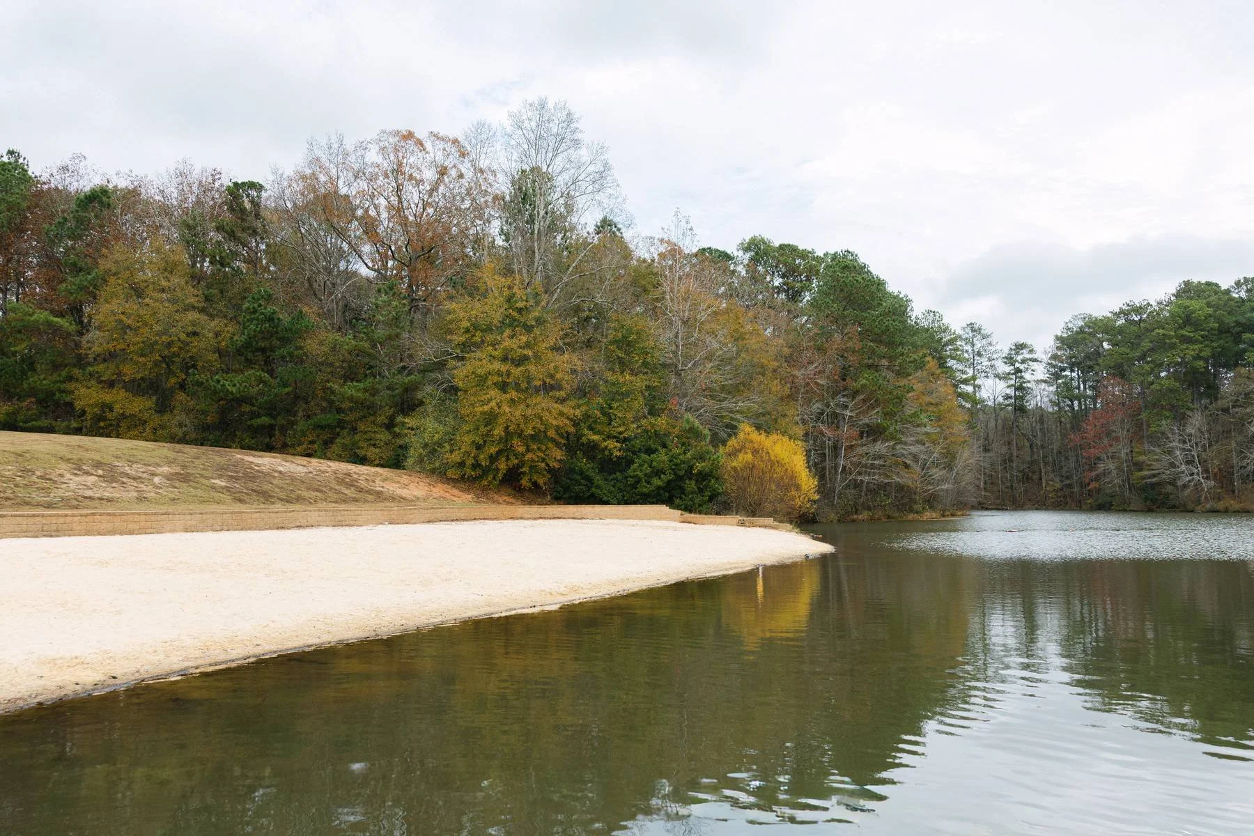 A shoreline beside a calm river with trees displaying fall foliage, some with green leaves, some with yellow and orange leaves, under a cloudy sky.