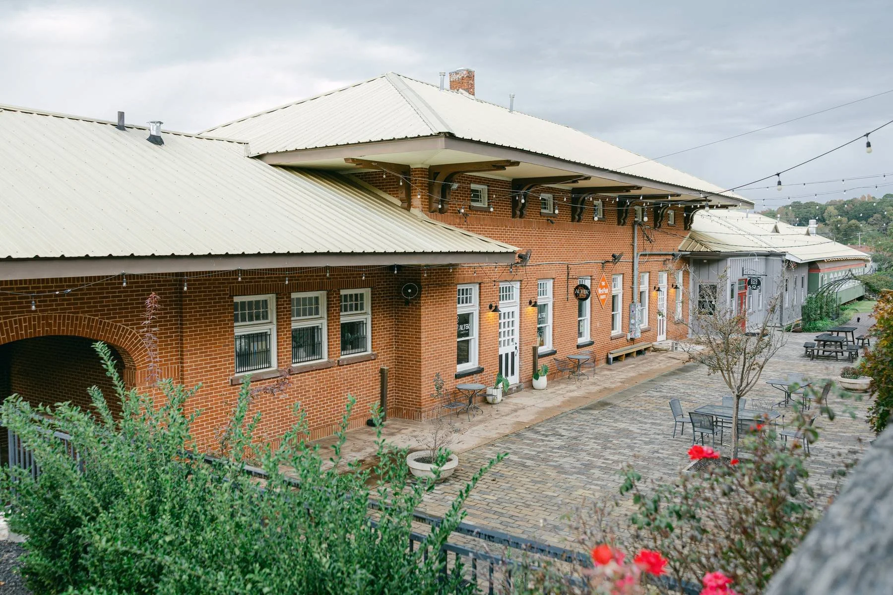 Outdoor view of a brick building with a yellow corrugated metal roof, string lights, benches, potted plants, and a small tree in a courtyard with cobblestone paving.