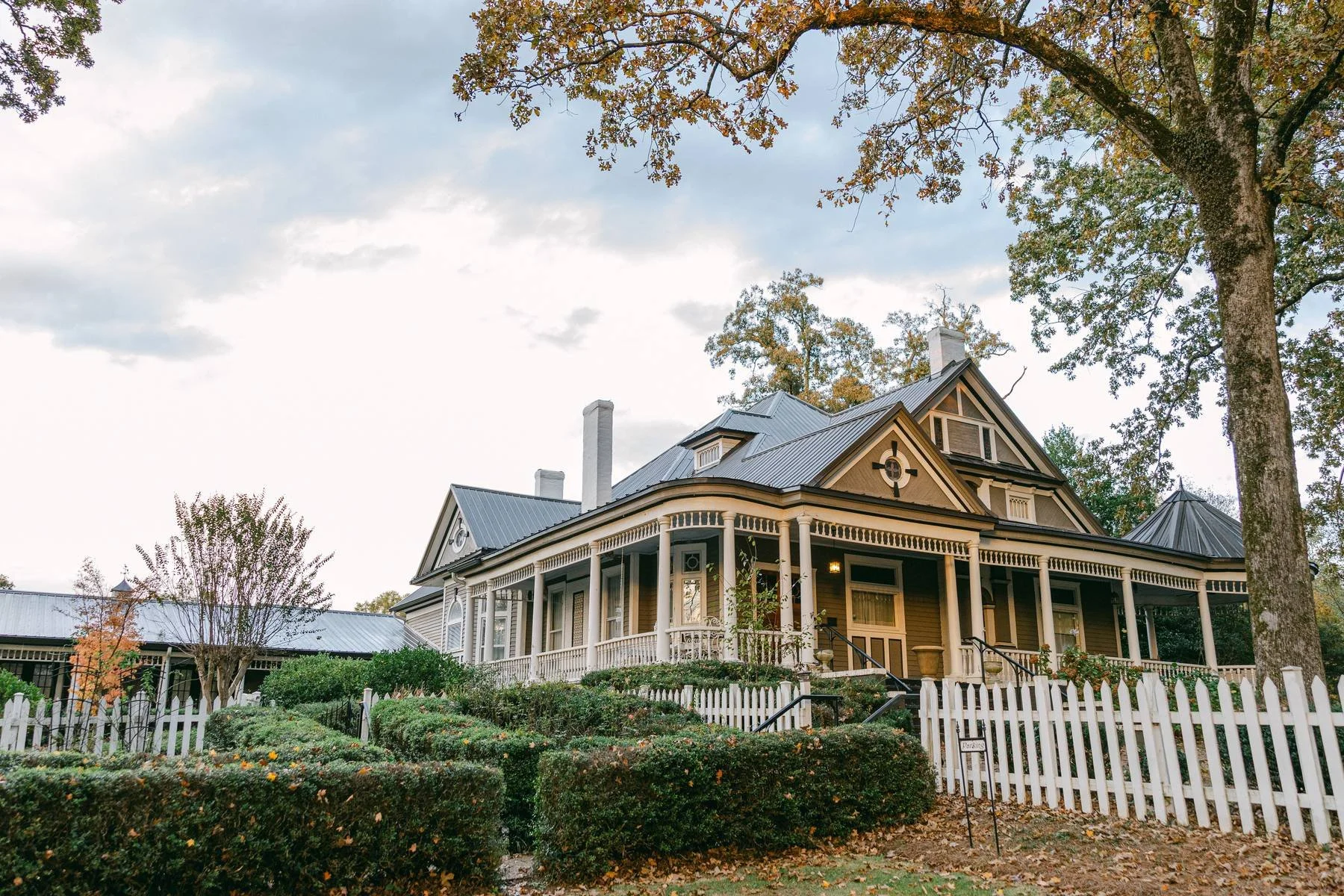 Victorian style house with a wraparound porch, white picket fence, surrounded by bushes and trees, under a cloudy sky.