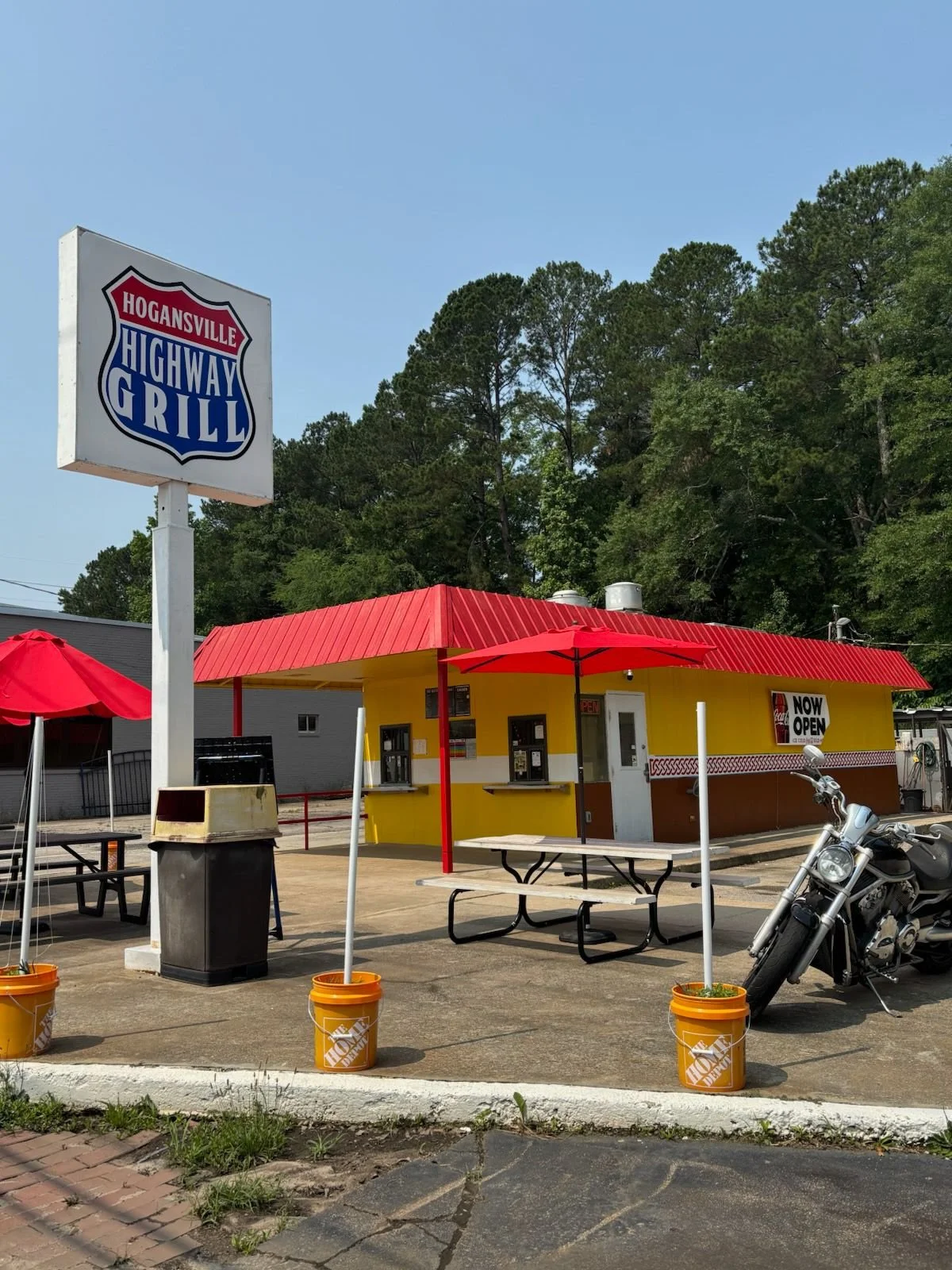A small yellow restaurant with red awnings and a red roof, outdoor seating with picnic tables, a motorcycle parked to the right, a "Now Open" sign, and a large sign with the words "Hogansville Highway Grill" in front of a wooded area.