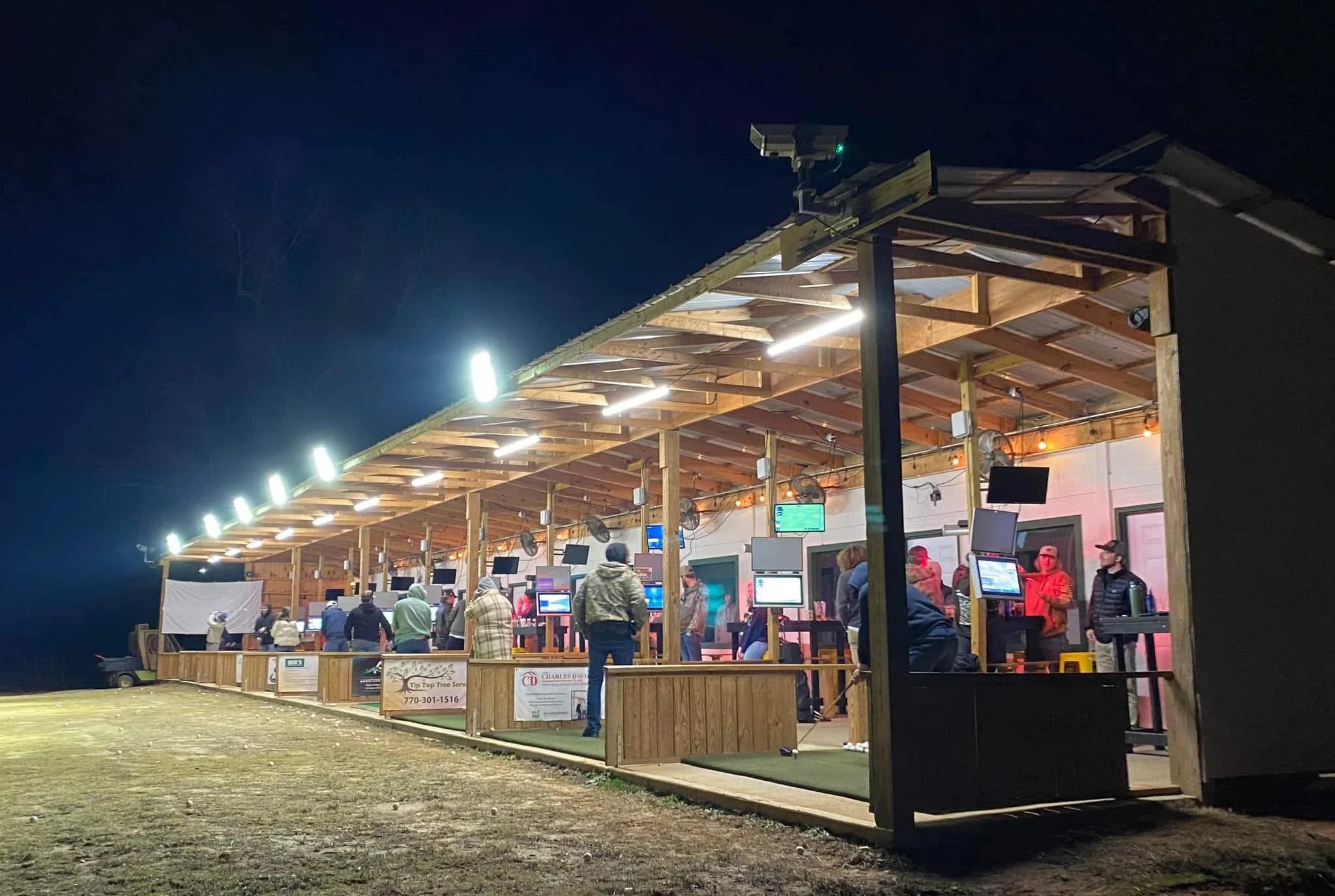 Night scene of a golf driving range with people practicing, illuminated by bright overhead lights and surrounded by a wooden structure.