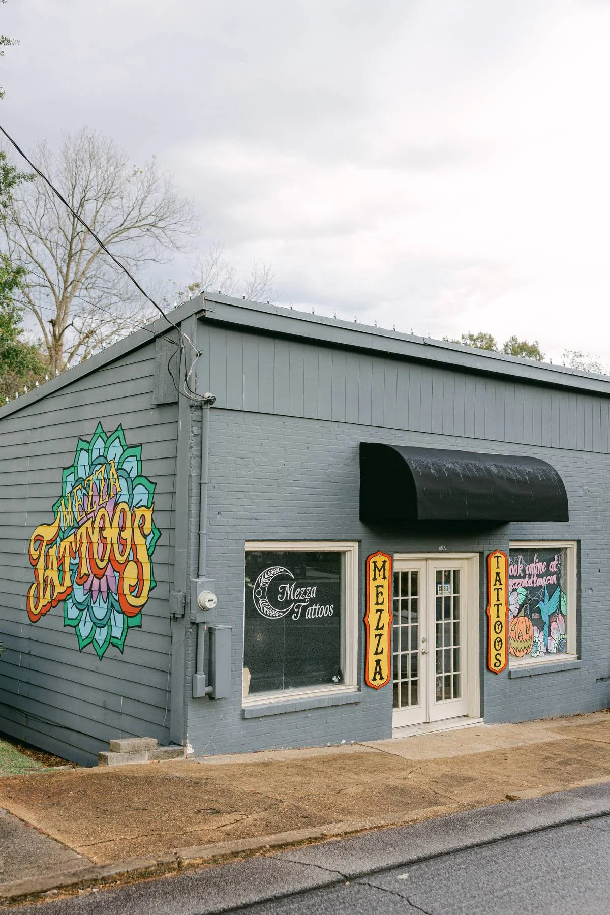 Gray building with colorful mural of a lotus flower on the side, black awning above the door, and windows displaying signs for 'Mezza Tattoos' and other services; concrete sidewalk in front.