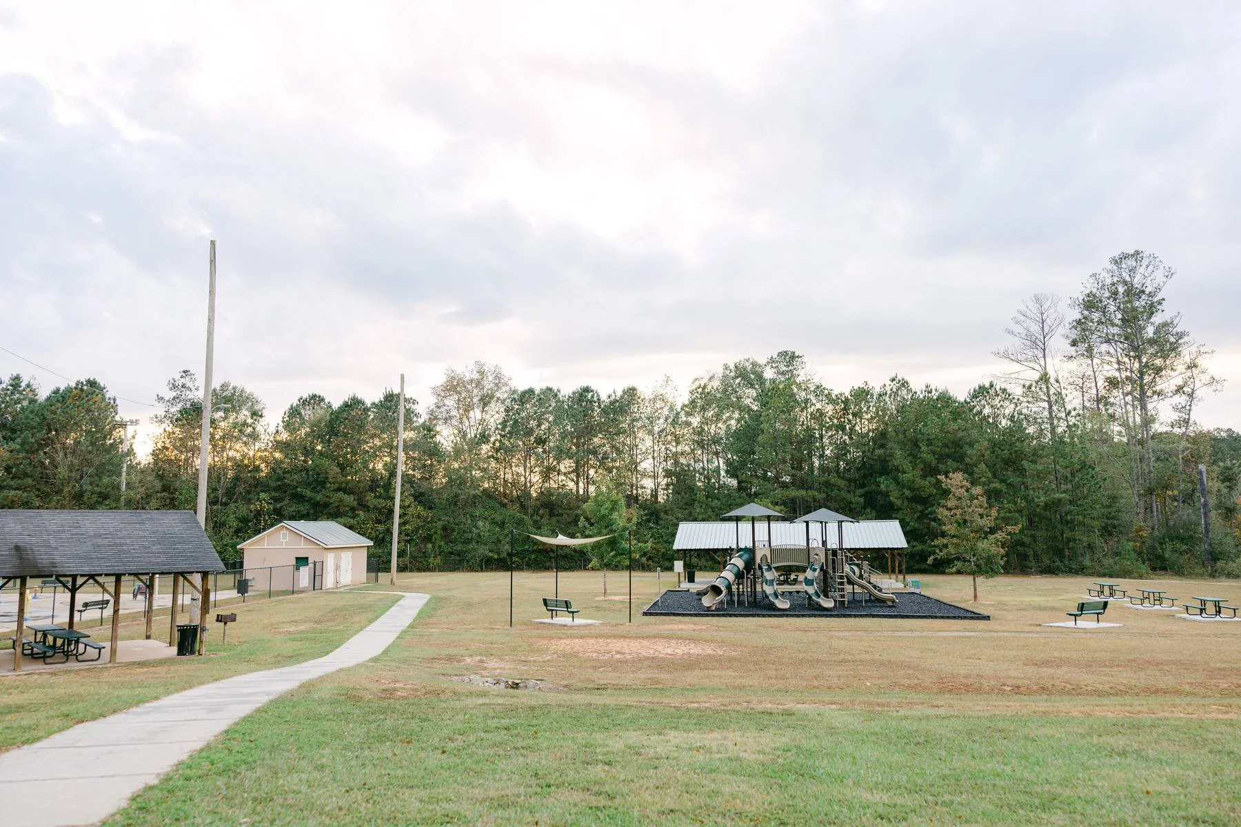 Empty playground with slides, picnic tables, a gazebo, and a path in a park surrounded by trees.
