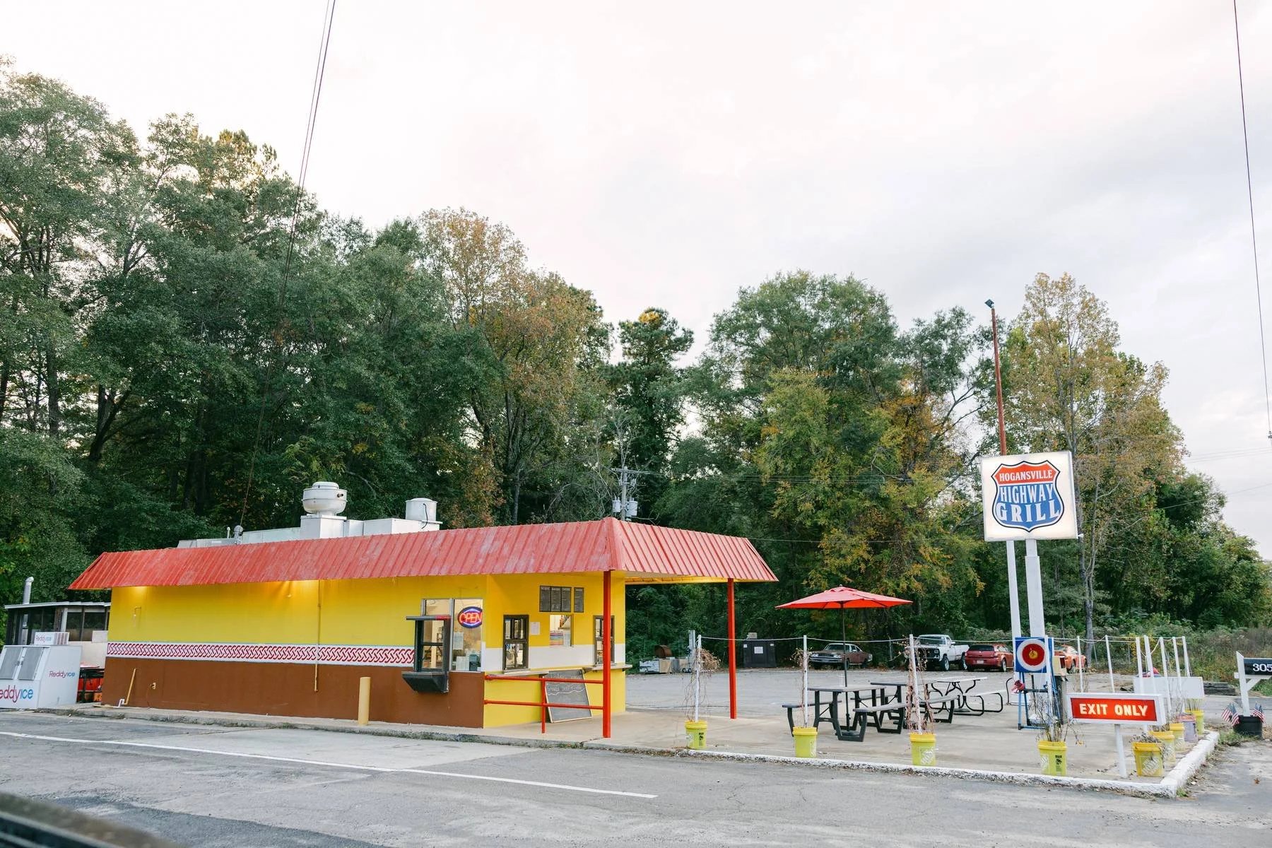 Colorful roadside restaurant with a red roof, yellow and brown walls, outdoor seating with picnic tables and a red umbrella, accompanied by a sign that reads "Hogansville Highway Grill."
