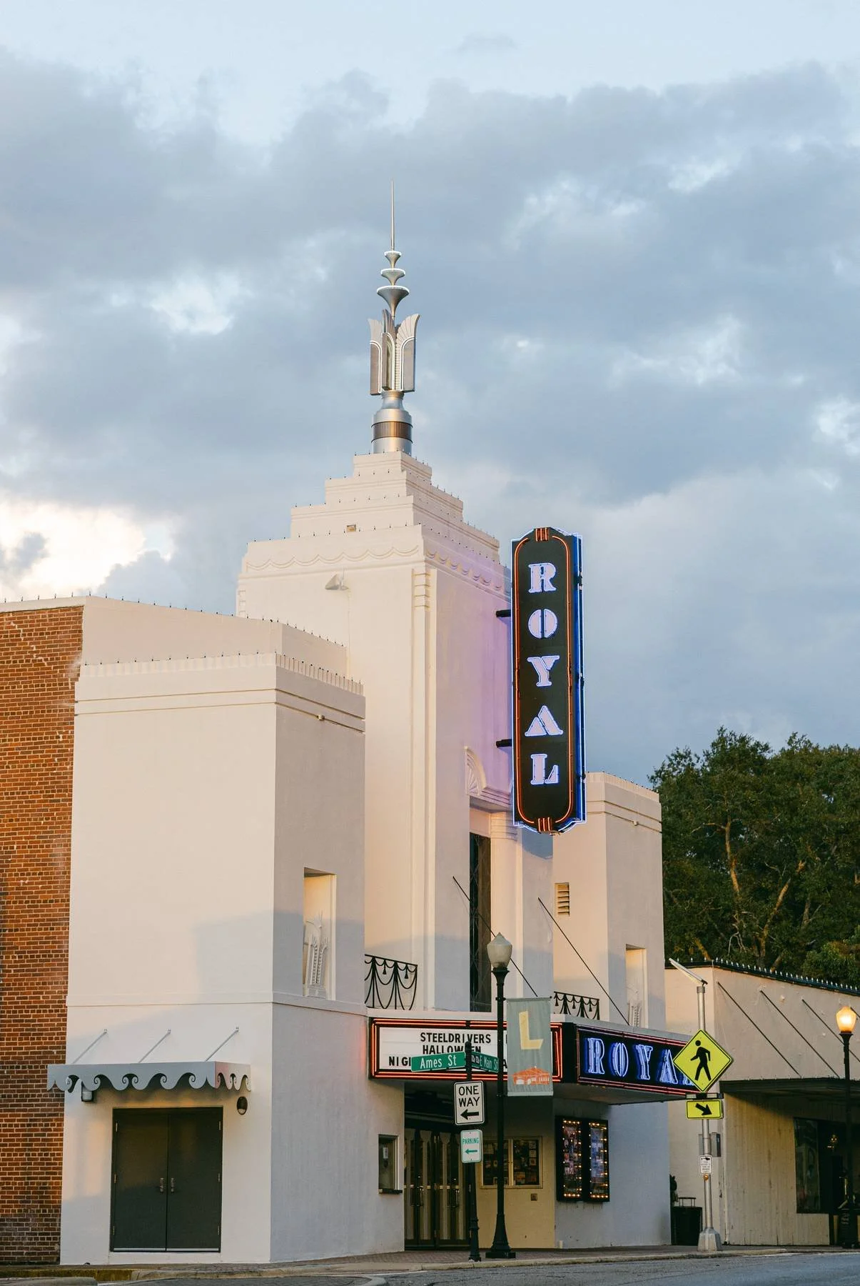 The Royall theater building with a vertical sign and a marquee, featuring a white facade, a tower with a spire, and surrounding street signs, including street names and a pedestrian crossing sign.