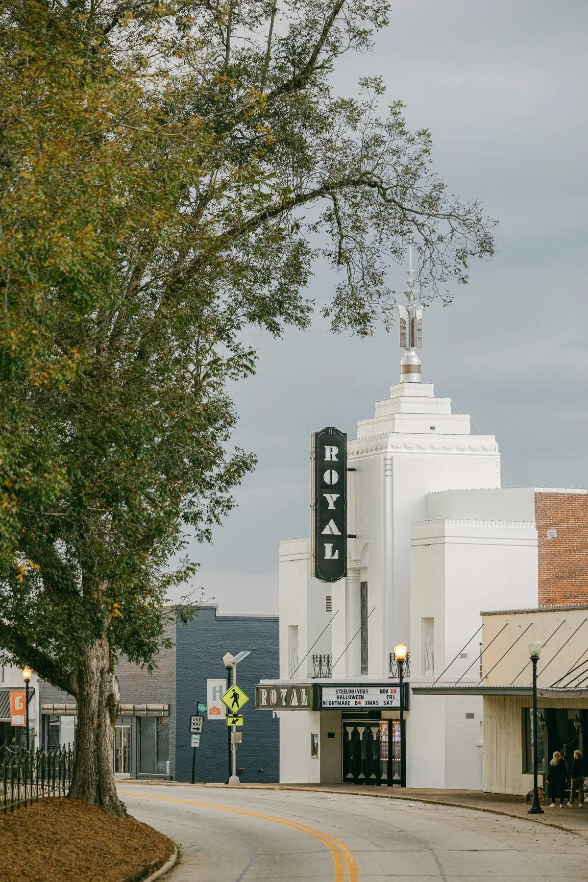 The image shows a street view with a large white building featuring a vertical sign that reads 'ROYAL' and a marquee displaying upcoming events. There are trees, street lamps, and pedestrians on the sidewalk, with a cloudy sky overhead.