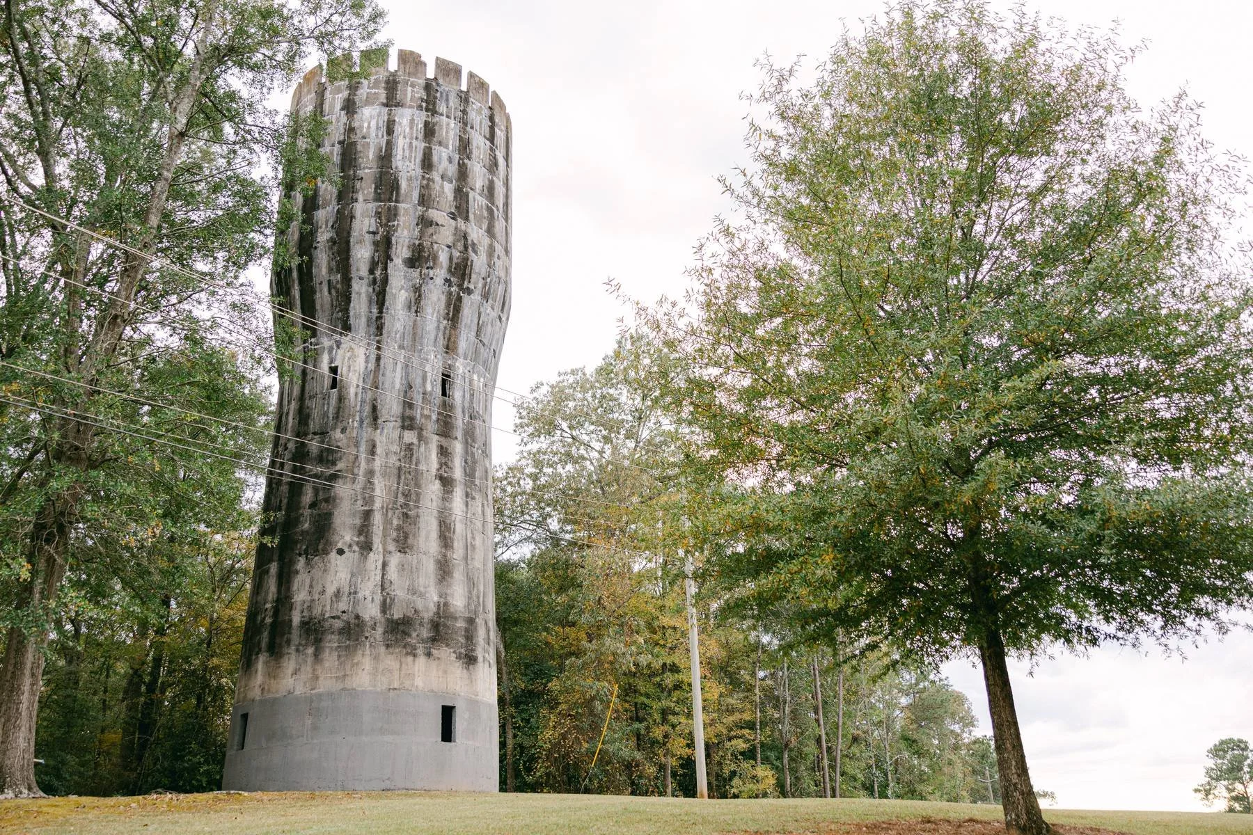 An old, weathered, tall concrete water tower surrounded by trees and a grassy area under a cloudy sky.