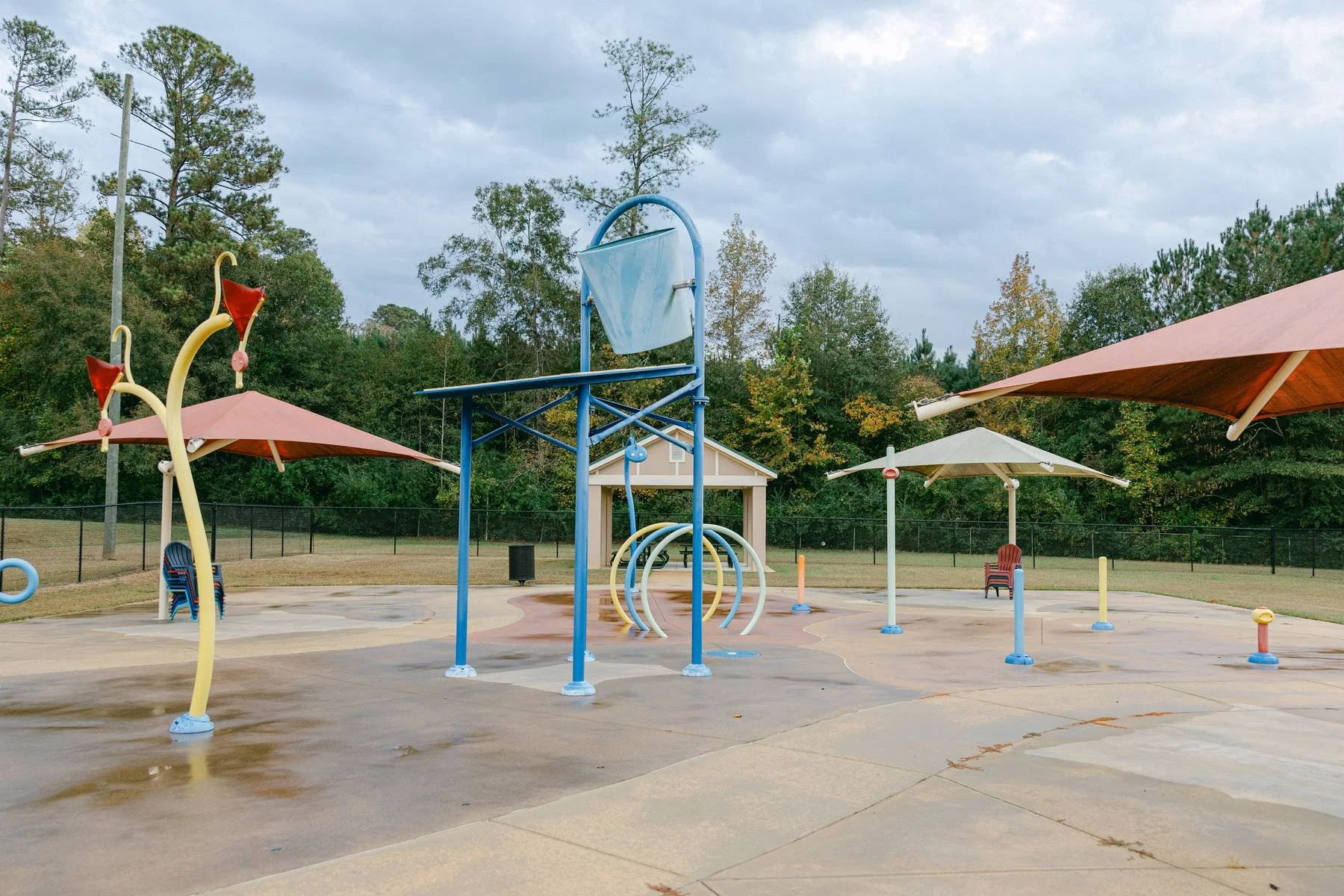 Empty water park with splash pads, fountains, and shaded seating areas with umbrellas, surrounded by trees and a cloudy sky.