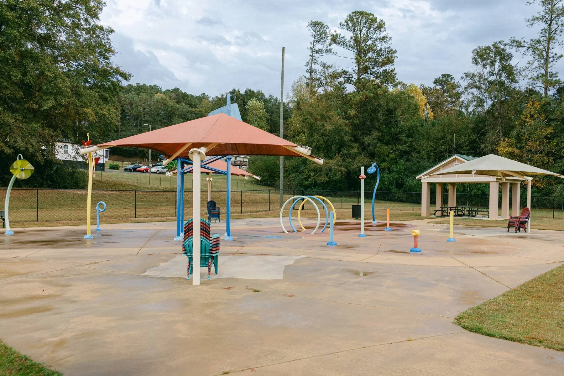 Empty colorful splash pad with umbrellas, climbing rings, and picnic pavilion, surrounded by trees.