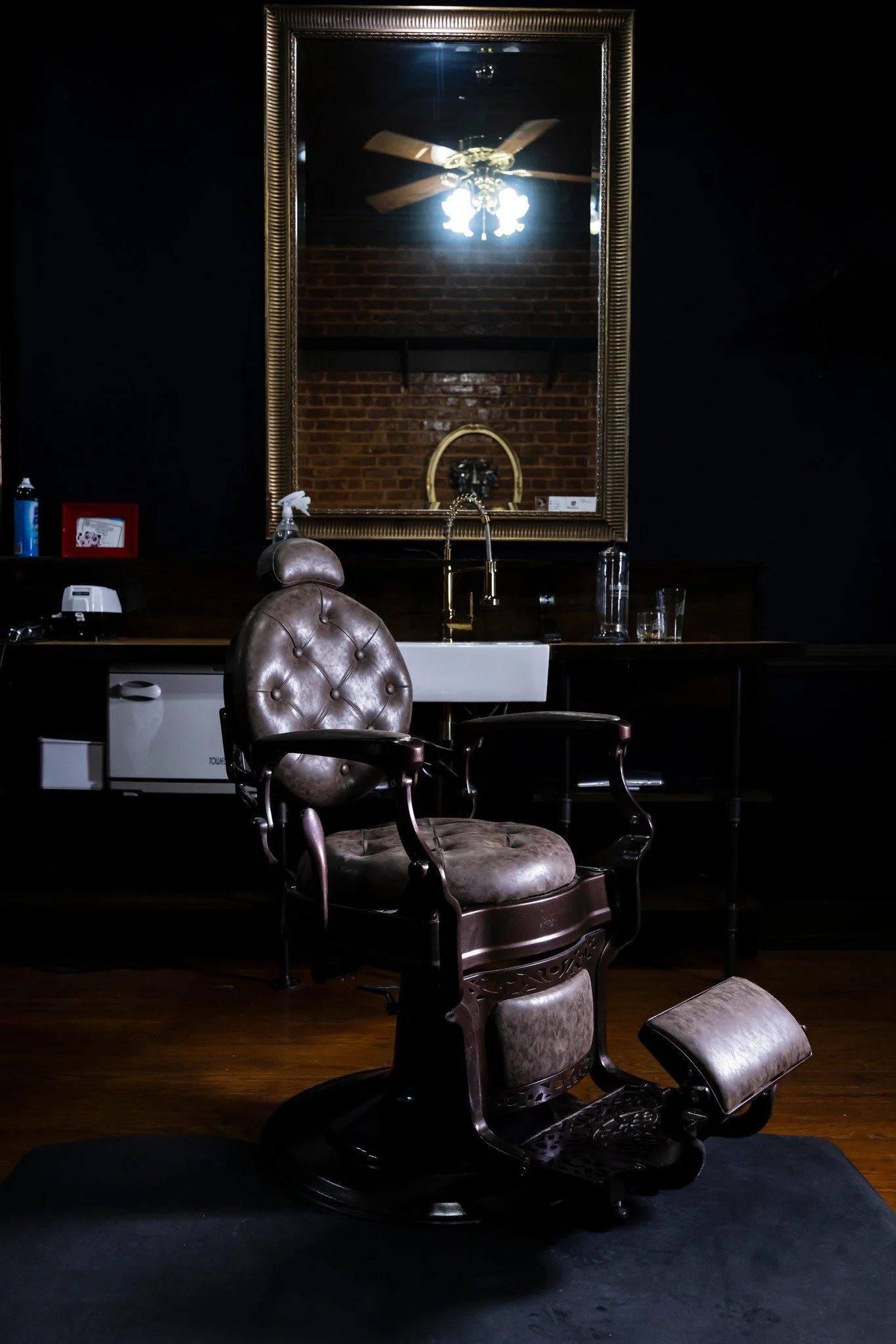 Vintage barber chair in front of a mirror with a ceiling fan reflection, dark interior with brick wall, gold faucet, and glasses on the counter.