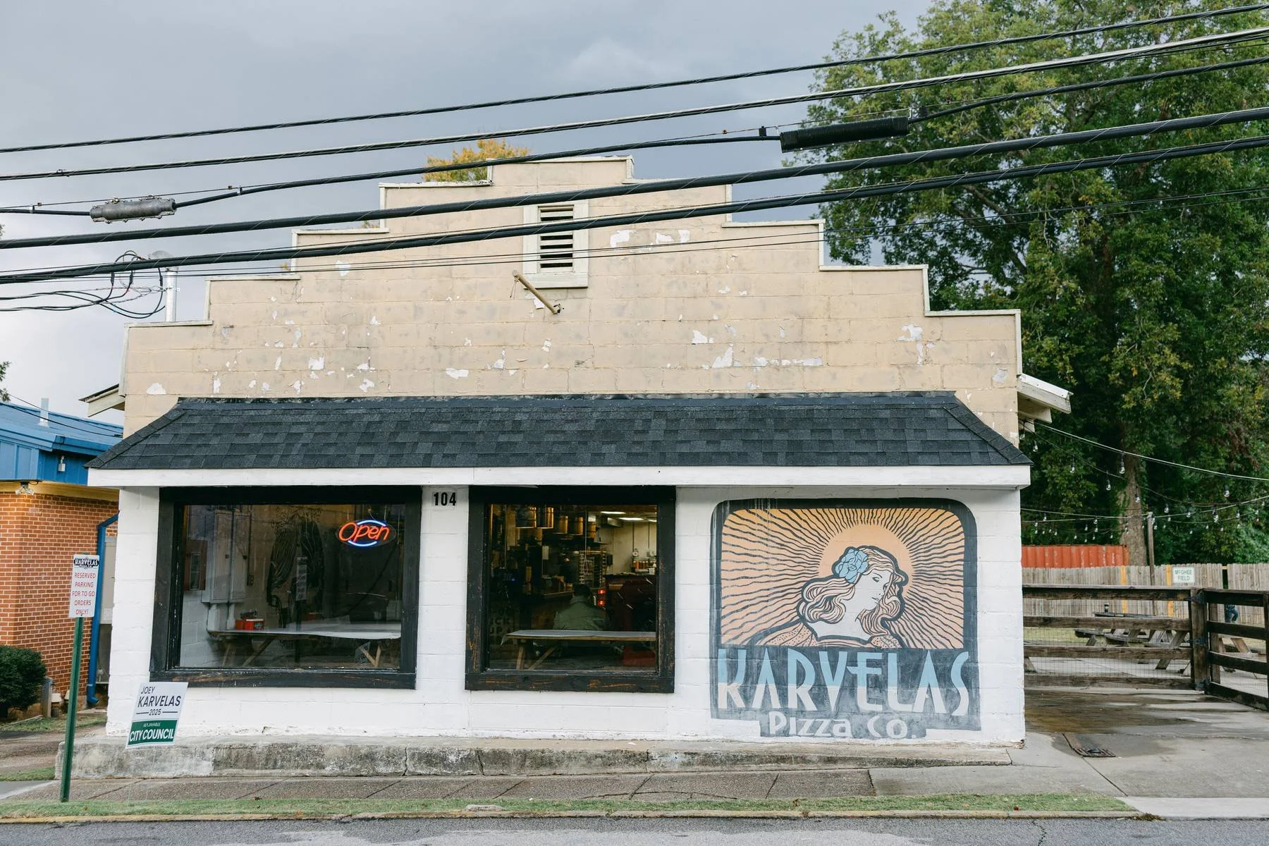 A small pizza restaurant with a hand-painted mural of a woman with a blue flower in her hair and the name 'Karvelas Pizza Co.' on the exterior wall. It has an neon 'Open' sign in the window, and a sign for city council election on the sidewalk.