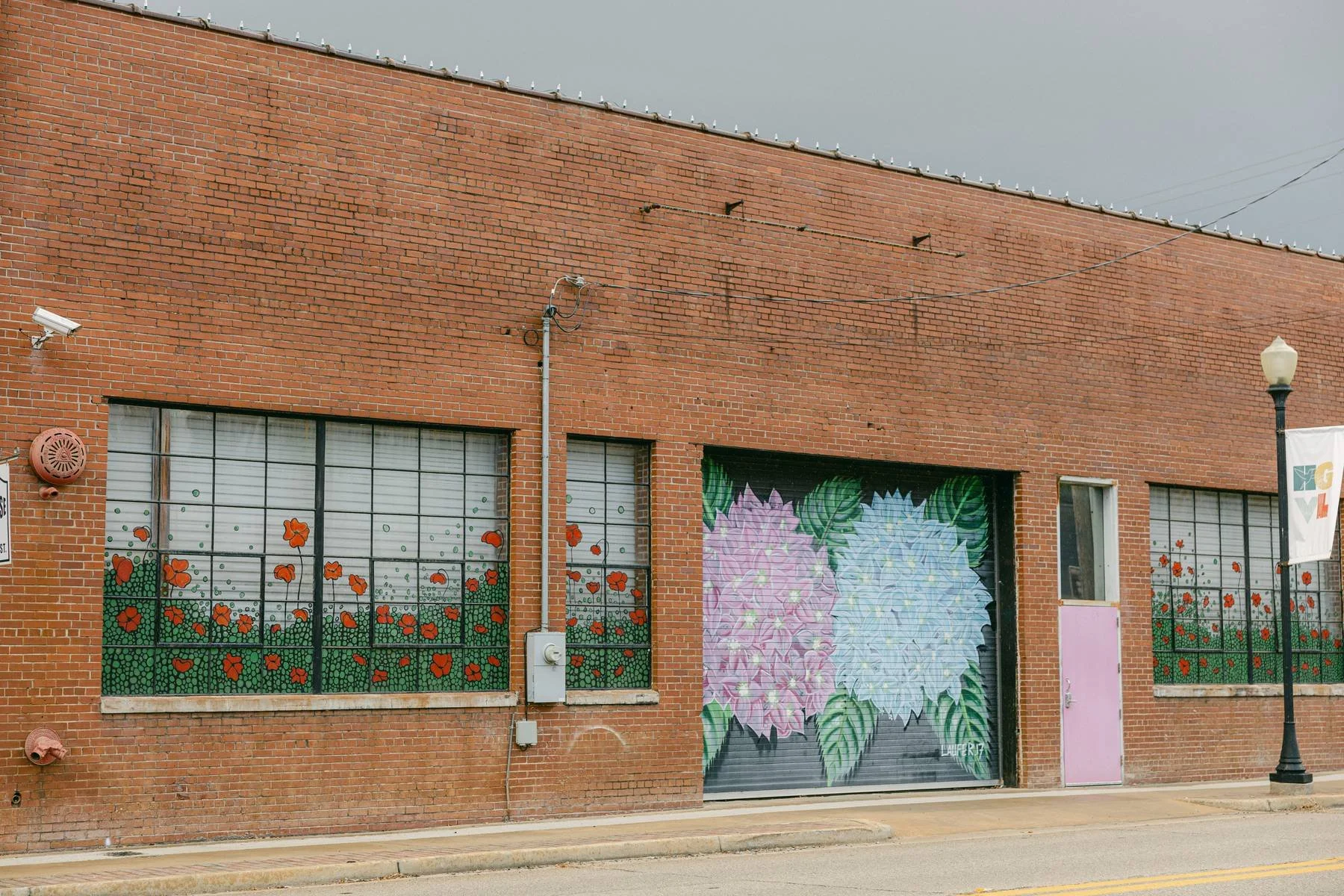 Street-level view of a brick building with large windows decorated with flower patterns and a mural of pink and blue flowers on a garage door, next to a pink door and a streetlamp.