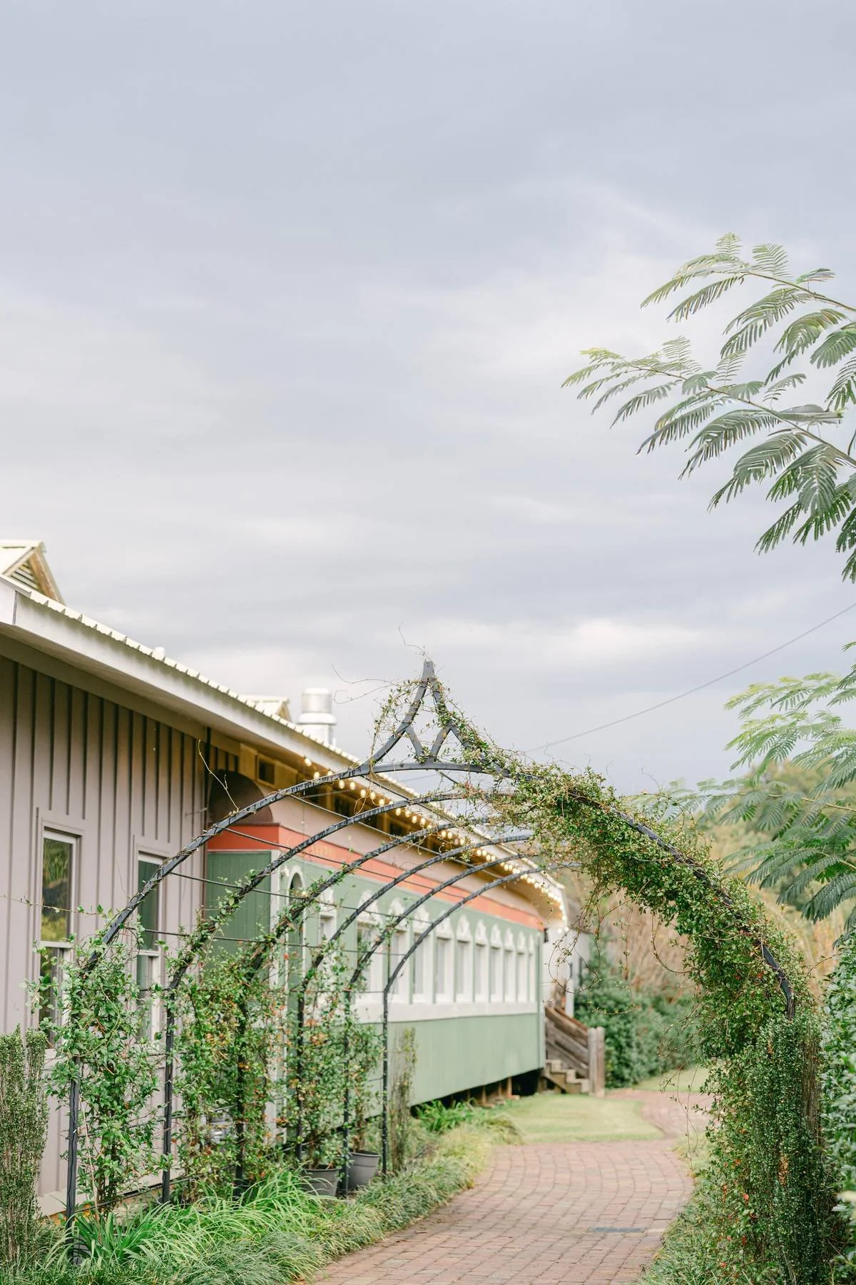 A decorative garden arch wrapped with greenery and fairy lights, situated along a brick pathway outside a house with beige siding and multiple windows, under a cloudy sky.