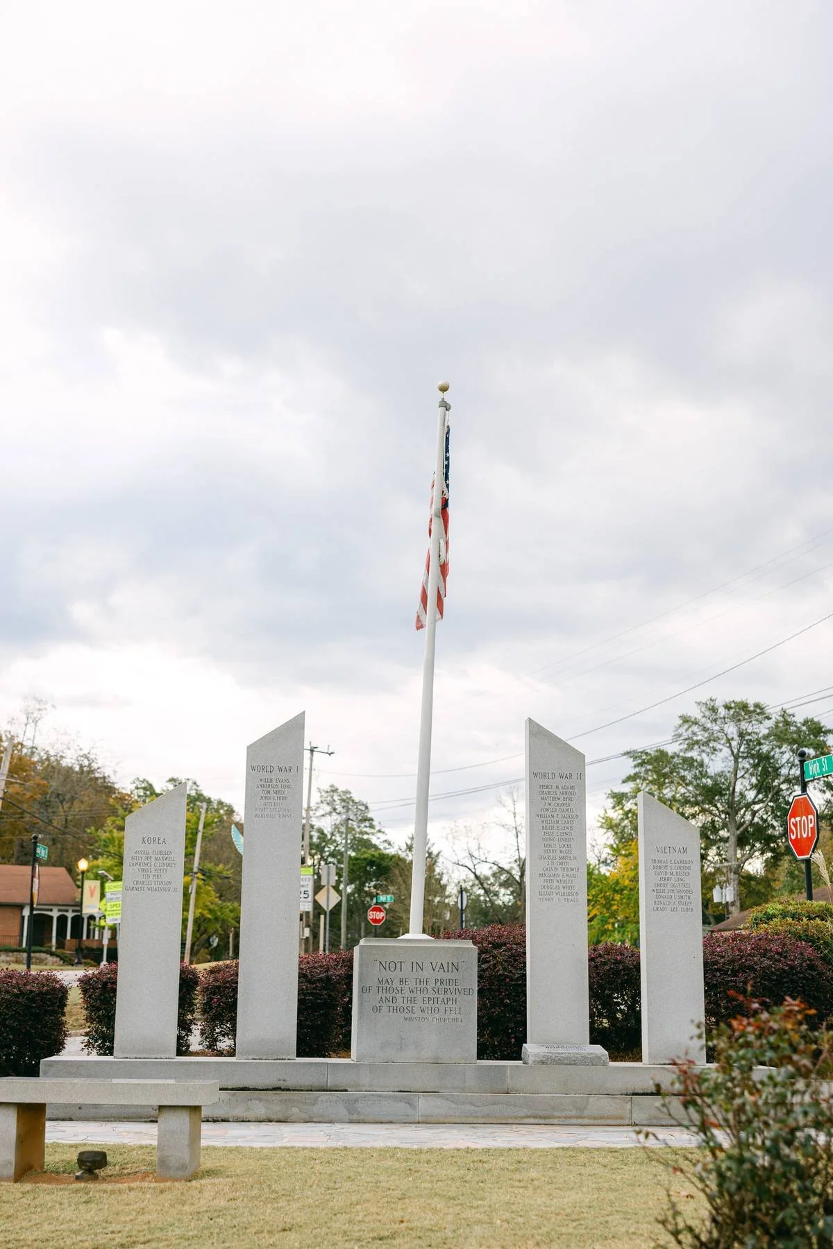 A memorial with five upright stone tablets inscribed with names of wars and soldiers, and a central plaque with an inscription by Winston Churchill. An American flag on a flagpole stands in front, with a cloudy sky above and trees and street signs in