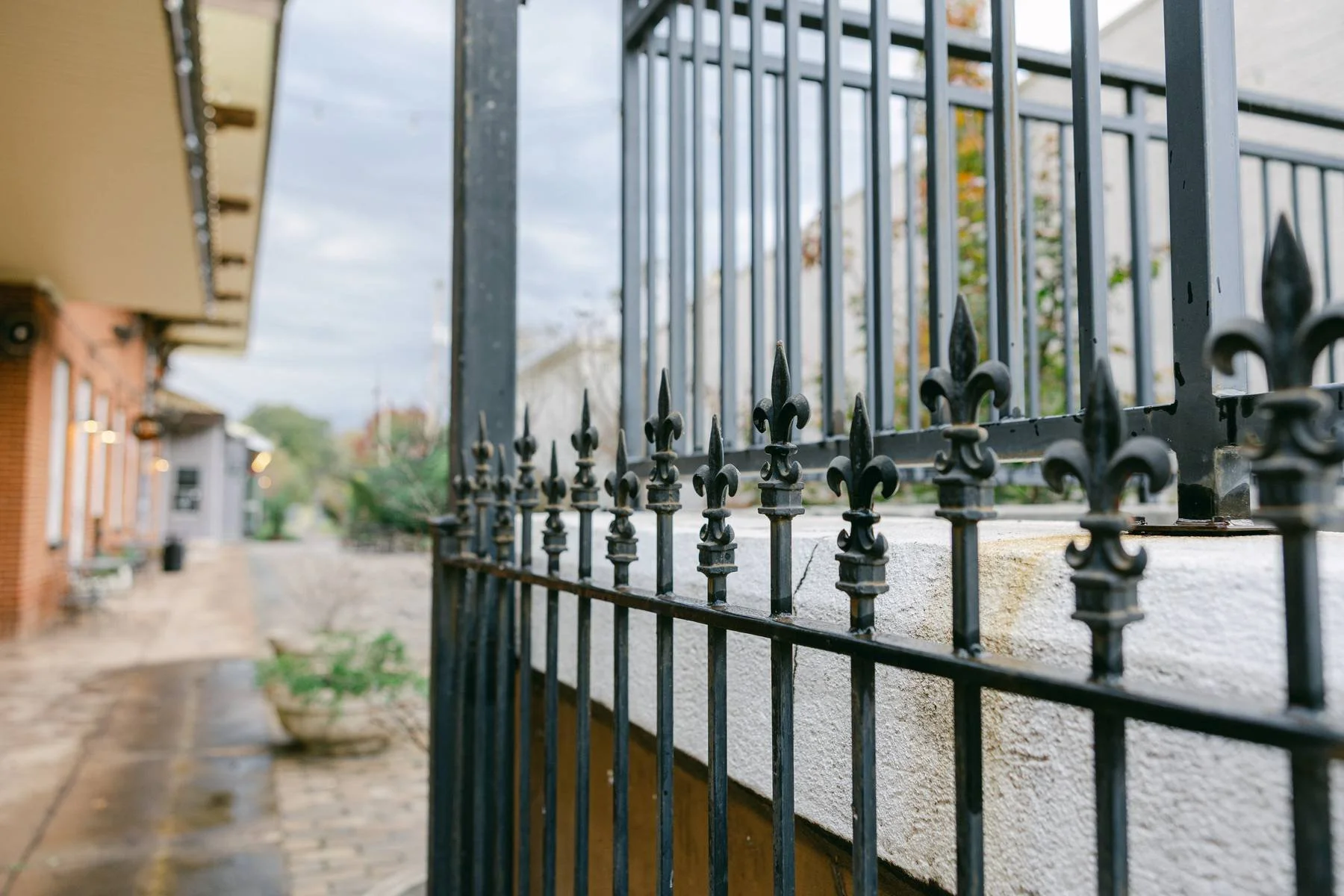 Close-up of a black wrought iron fence with decorative fleur-de-lis finials, mounted on a white concrete wall, along a sidewalk on a cloudy day.