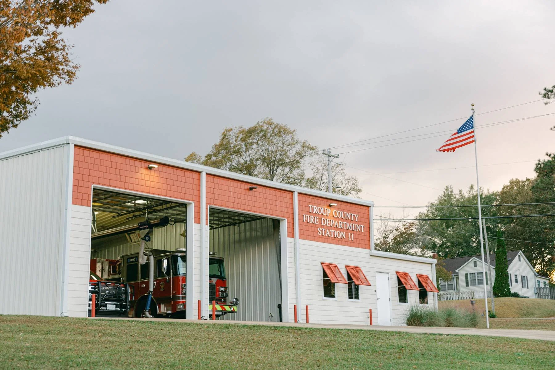 Fire station building with red and white exterior, American flag flying nearby, trucks visible inside the open garage, located in a suburban area with trees and houses.
