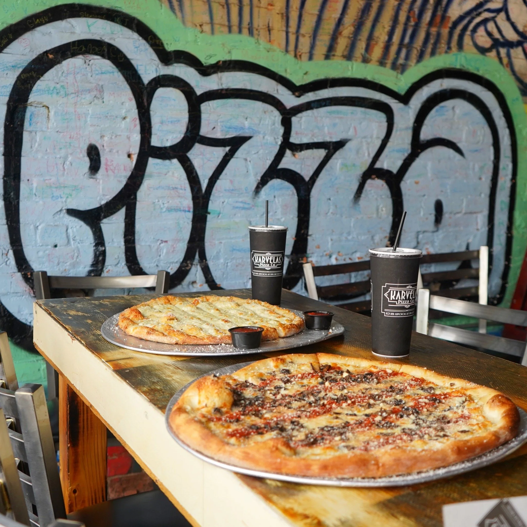 Two pizzas, one with cheese and the other with toppings, on metallic trays on a wooden table. Two black drinks with straws and the logo 'Karvelas Pizza Co.' are also on the table. In the background, there is a colorful graffiti wall with the word 'PEACE' in large letters.