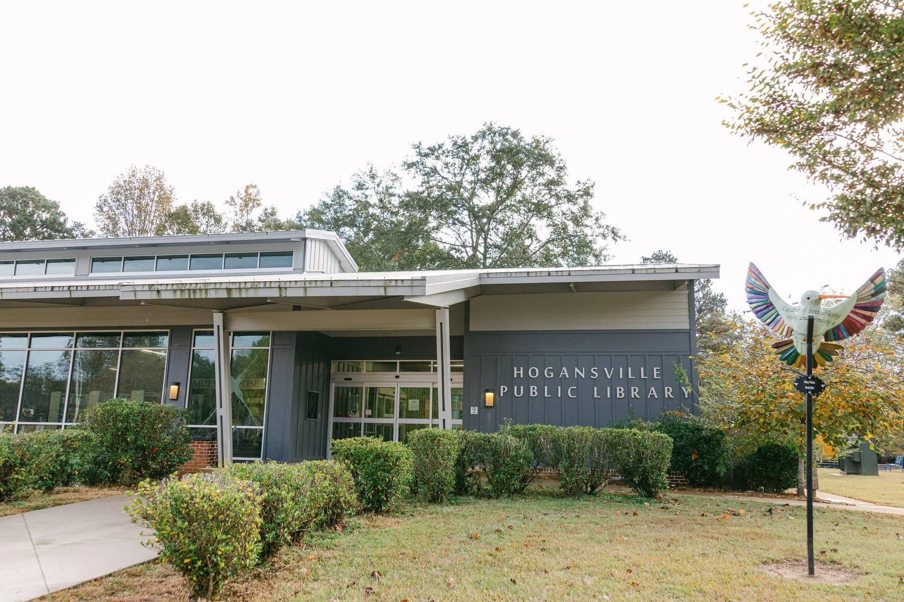 Exterior of Hogansville Public Library with bushes, trees, and a colorful bird sculpture in front.