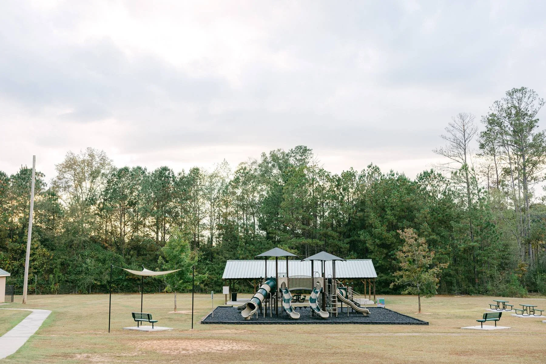 Empty playground with slides, tunnels, and swings, surrounded by trees and picnic benches on a cloudy day.