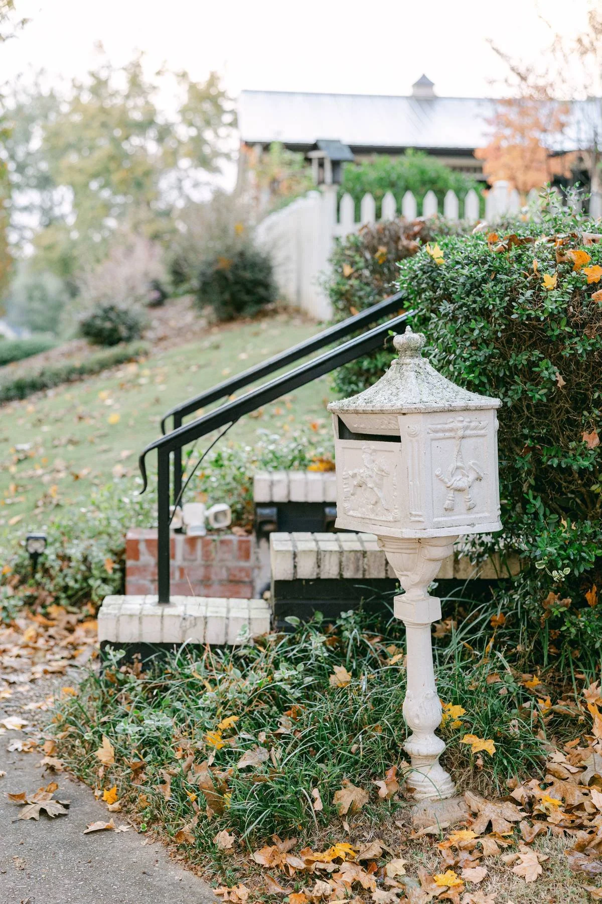 A white decorative mailbox with lion carvings, standing on a pedestal, beside a garden hedge and a black railing near brick steps leading up a lawn, with fall leaves on the ground.
