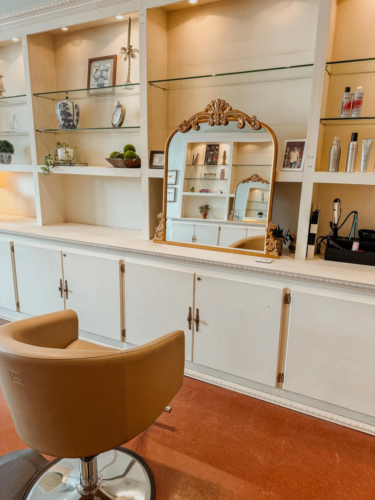 A salon workstation featuring a large mirror with an ornate gold frame, wooden cabinets and shelves displaying decorative items and hair products, a tan salon chair, and a red-brown floor.