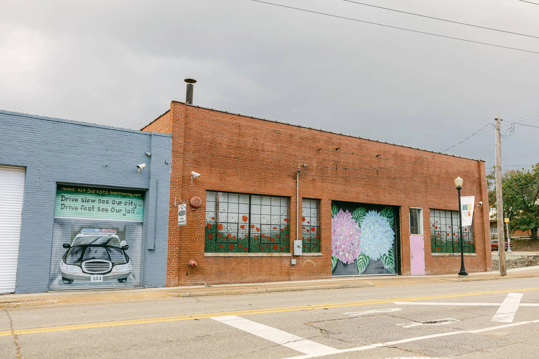 A brick building with large windows decorated with painted flowers, next to a blue building with a mural of a police car and text about driving and jail, on a street with a lamp post and a power pole.