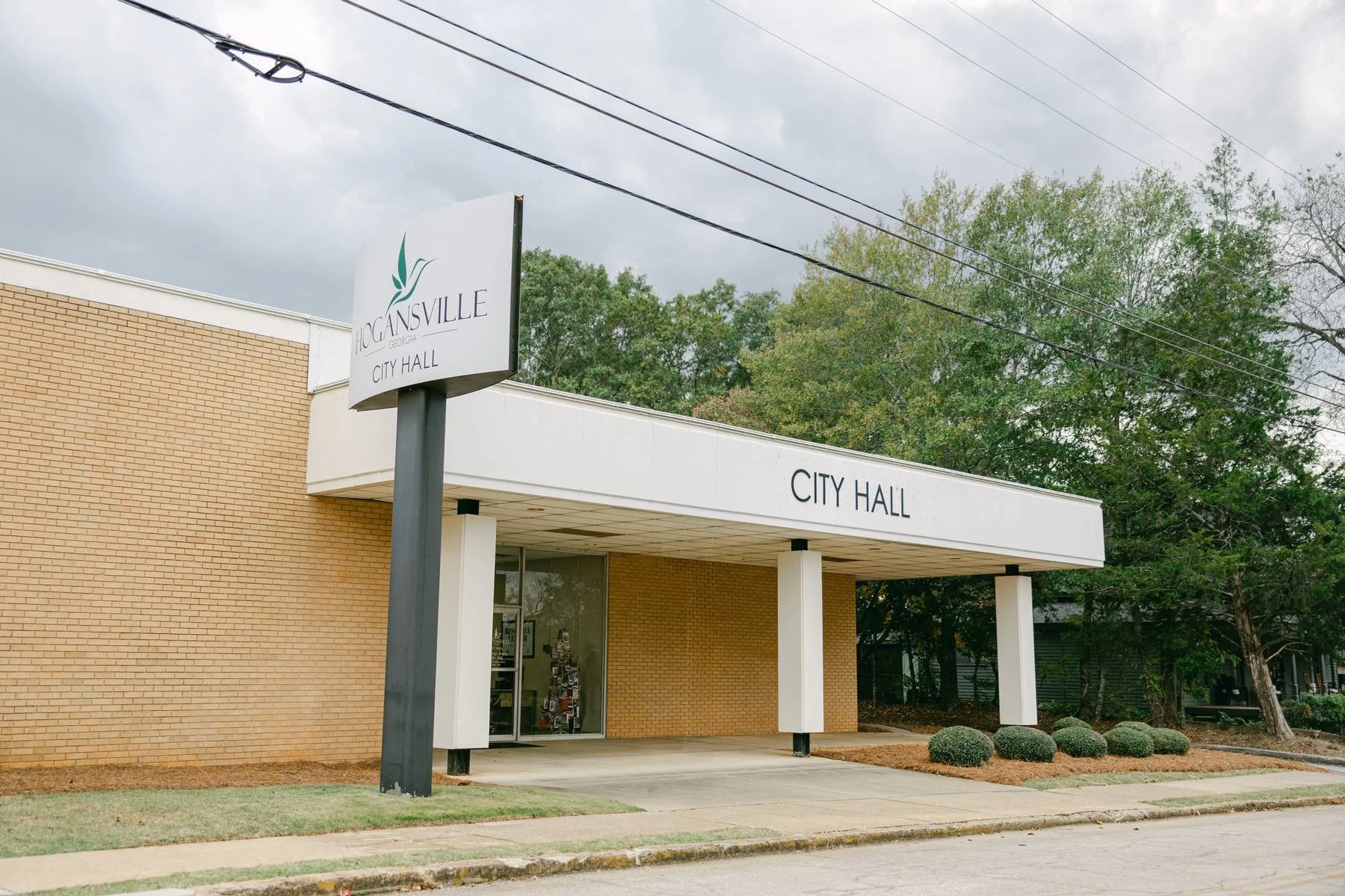 Exterior of Hogansville City Hall building with brick walls and a white awning. Sign on a pole in front reads 'Hogansville Georgia City Hall'.