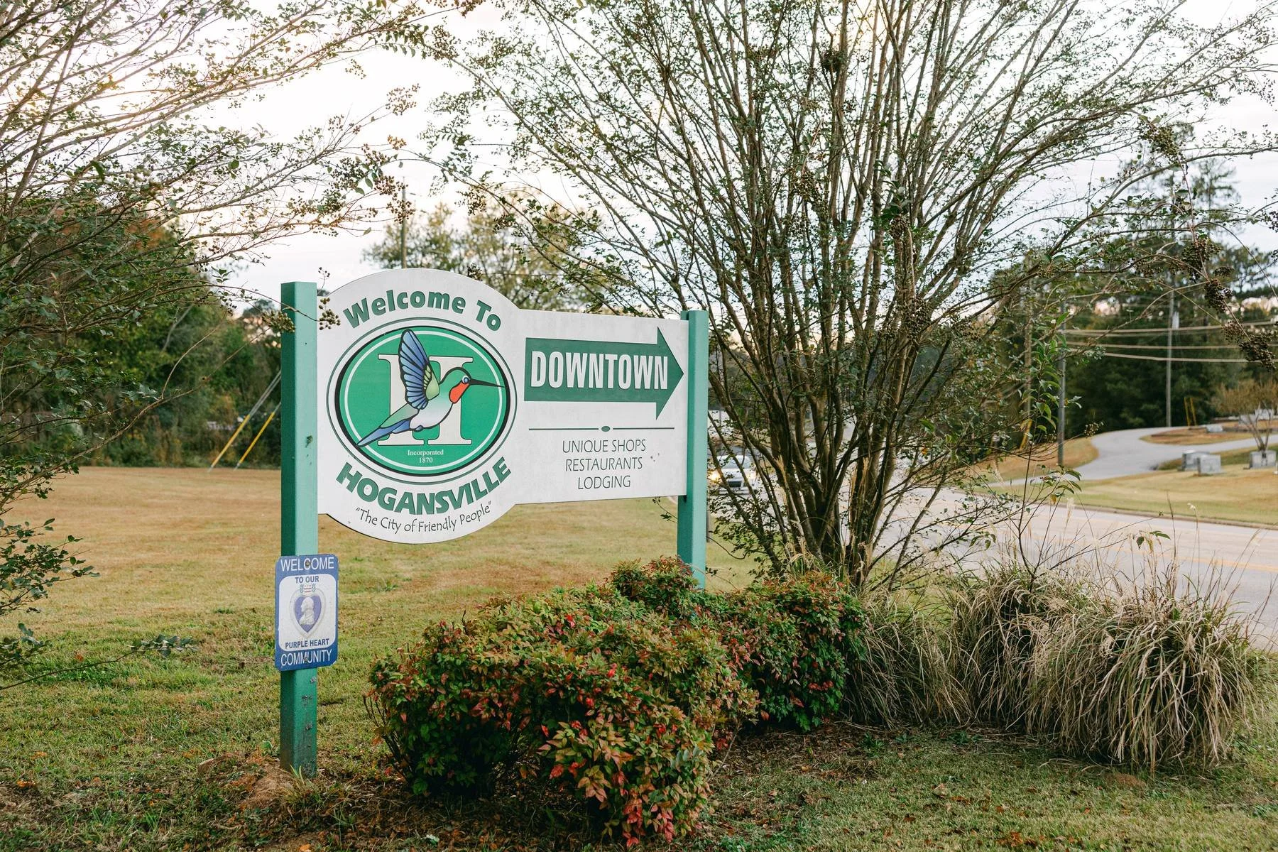 Sign welcoming visitors to Hogansville, Georgia, with directions to downtown, featuring a hummingbird logo and mentioning shops, restaurants, and lodging.