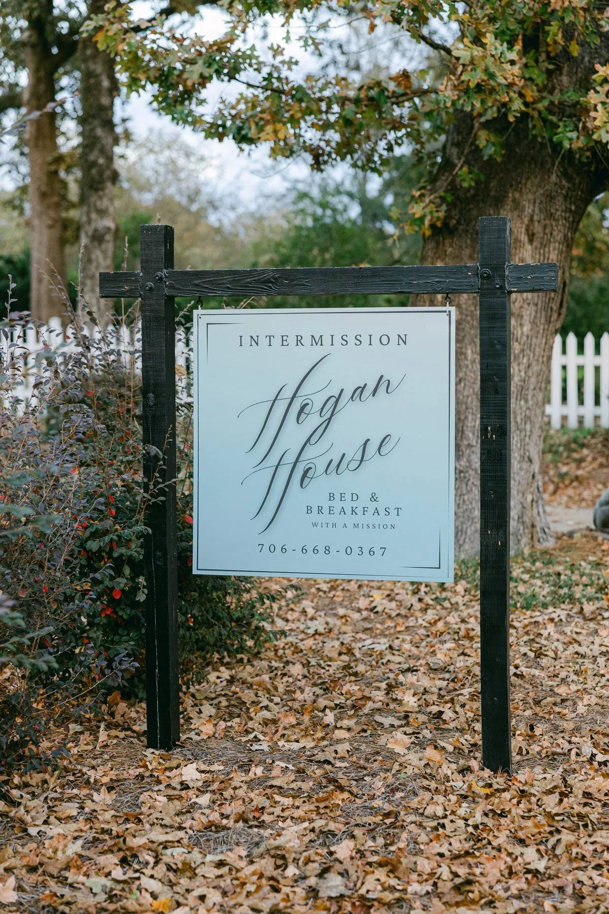 Sign reading 'Intermission Hogan House Bed & Breakfast with a mission, 706-668-0367' in front of tree and bushes with fallen leaves on ground.