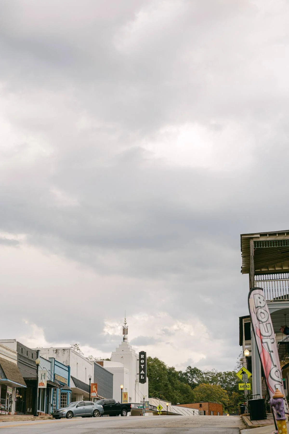A street scene in a small town with storefronts and cars parked along the road, historic architecture, and a tall structure with a spiritual symbol in the distance under cloudy skies.
