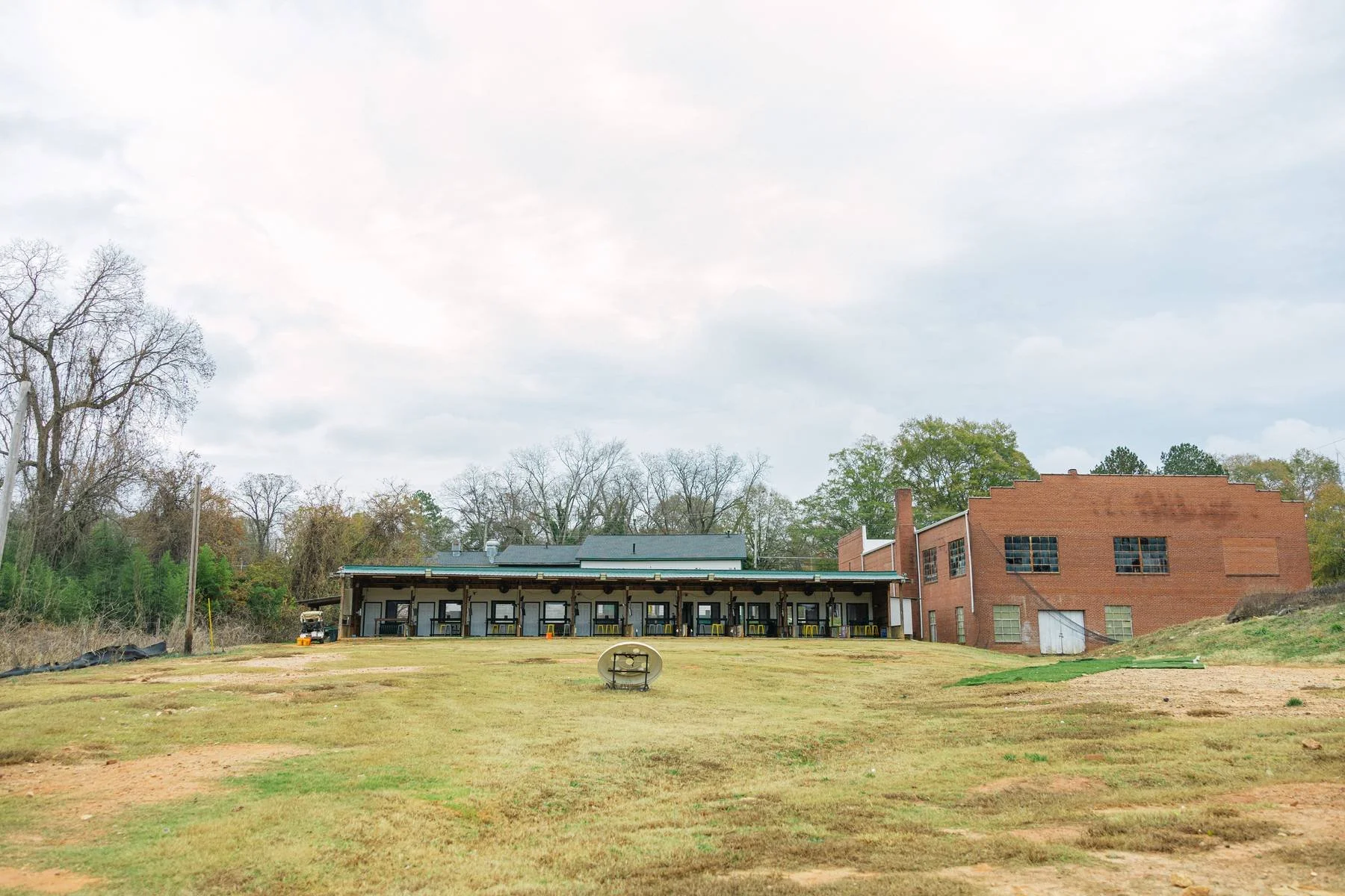 Empty grassy lot in front of a building with a covered porch and multiple doors, a discarded toilet on the lawn, and a red brick structure to the right, under cloudy sky.