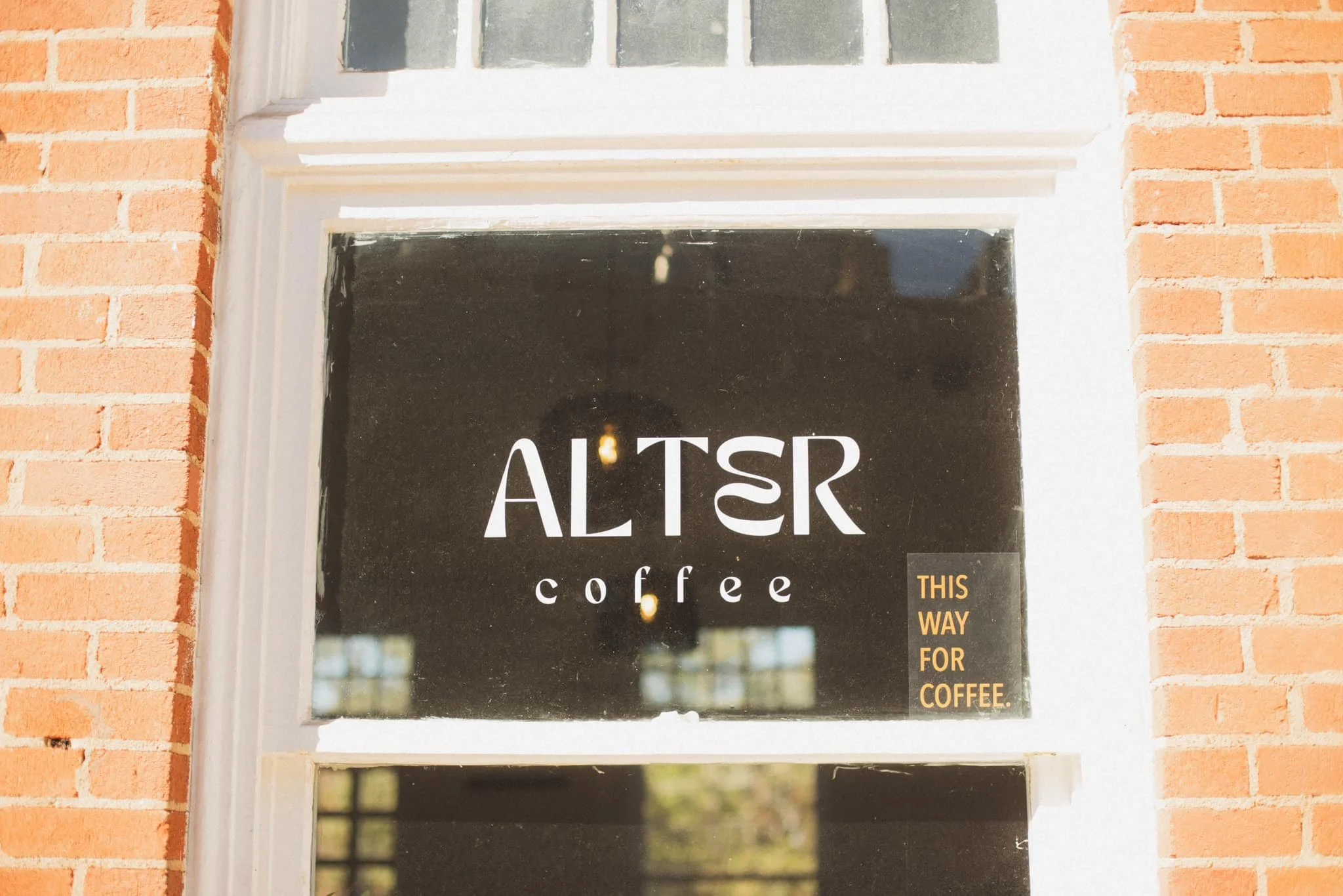 A black sign with white text that reads 'ALTER coffee' and a smaller, yellow text box that says 'THIS WAY FOR COFFEE.' The sign is on a brick wall with a white window frame around it.