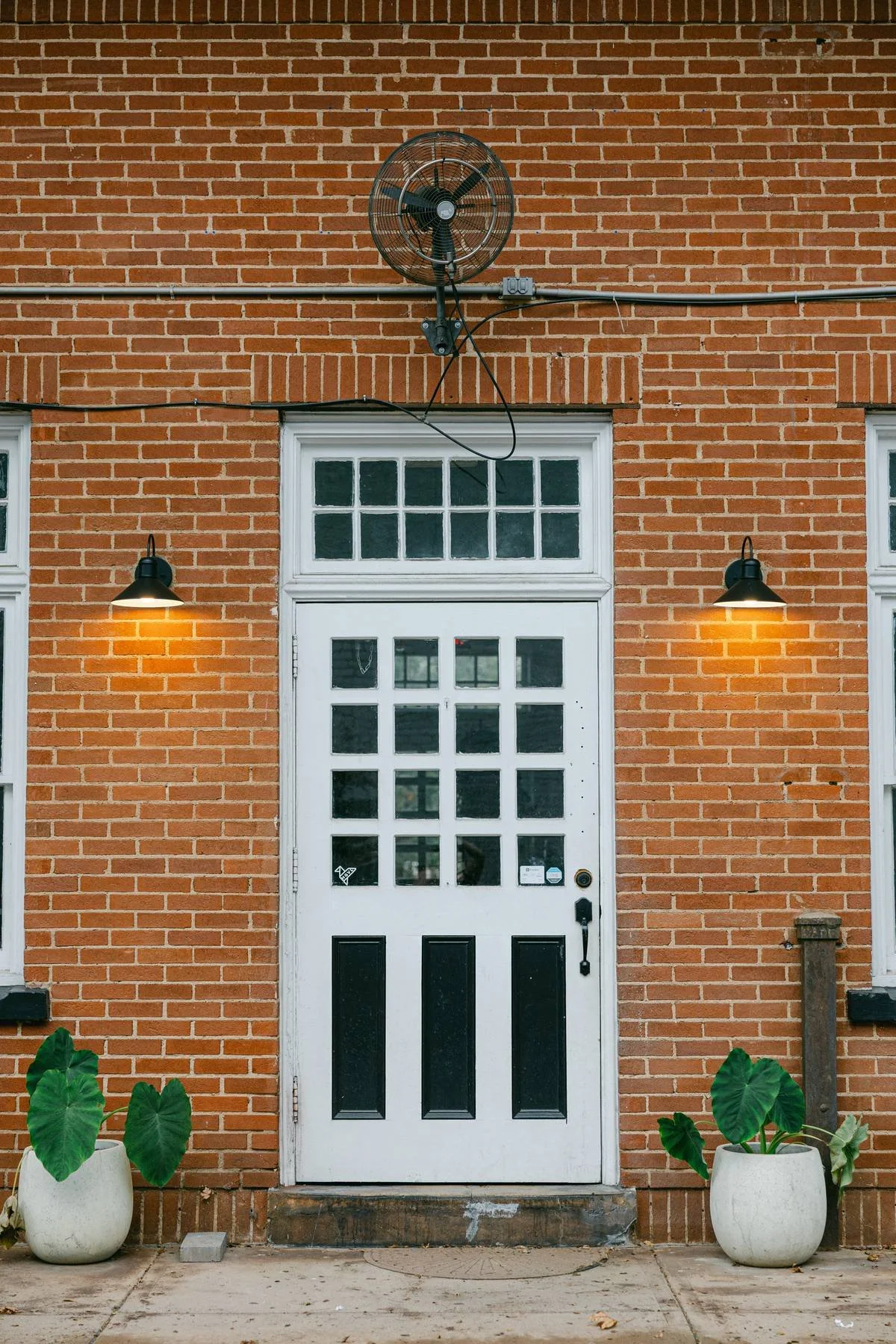 Brick building with front door, two potted plants, wall-mounted lamps, and a mounted fan above the door.