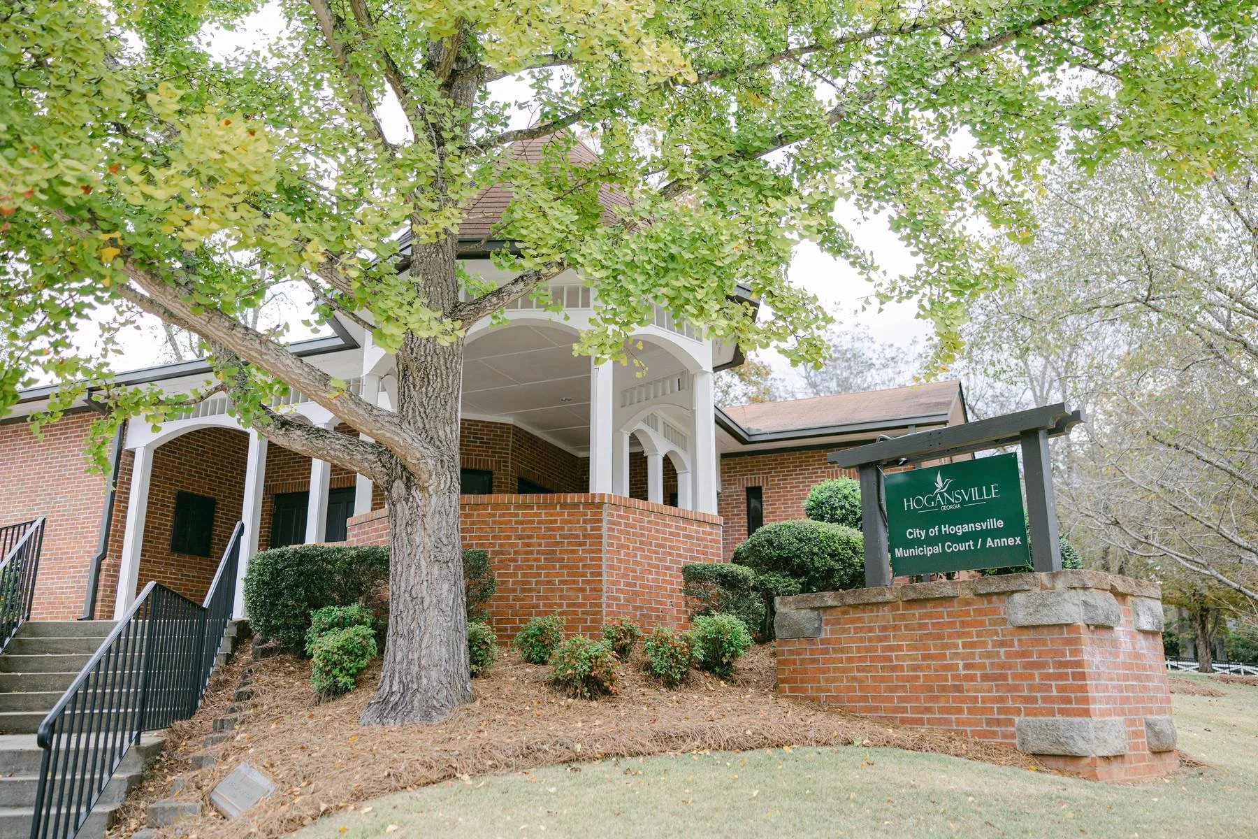 The exterior of the City of Hogansville Municipal Court / Annex building with a brick facade, surrounded by bushes, a tree, and a sign indicating its location in Hogansville, Georgia.