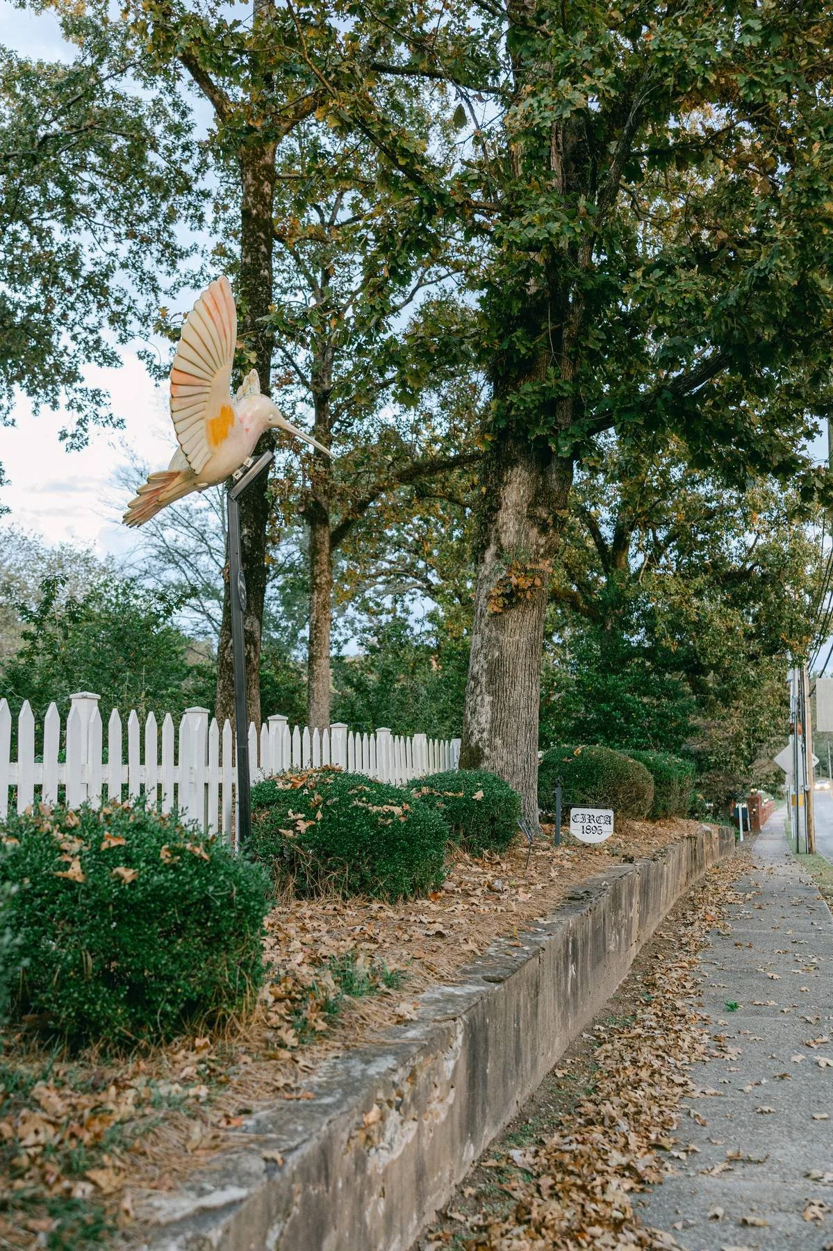 A decorative sign of a hummingbird mounted on a pole near a sidewalk, with trees, bushes, a white picket fence, and fallen autumn leaves in the background.