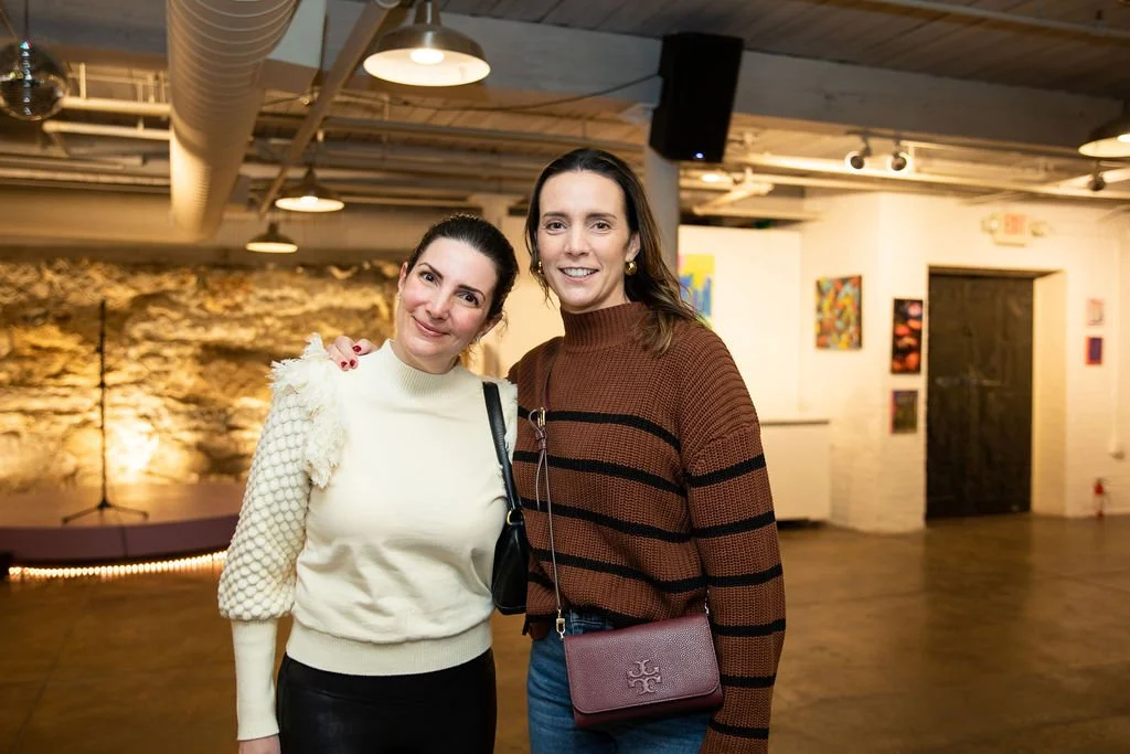 Two women in a warmly lit indoor space, one wearing a white sweater and the other a brown and black striped sweater with a maroon bag. Background includes art on white walls and a stone wall.