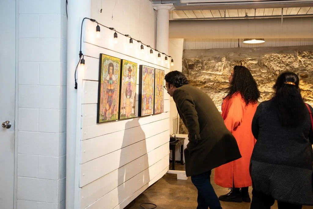 Three people viewing colorful paintings in a gallery with exposed brick walls and overhead lighting.